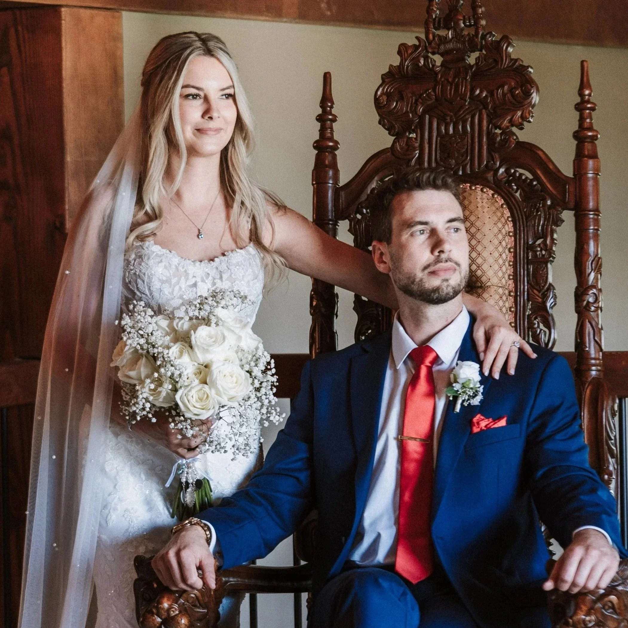 A bride with long blonde hair in a wedding dress holding a bouquet of white roses and baby's breath, standing next to a groom with dark hair and beard in a blue suit with white shirt and red tie, seated on an ornate wooden throne-like chair.