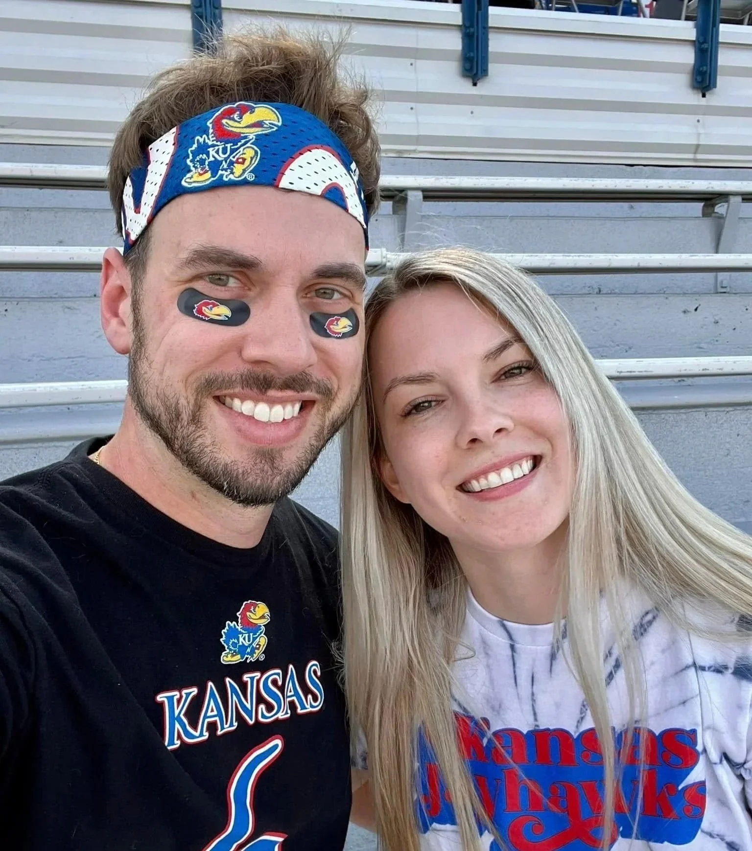 A group of Kansas Jayhawks fans at a stadium, wearing team shirts and face paint, watching a game on the field.