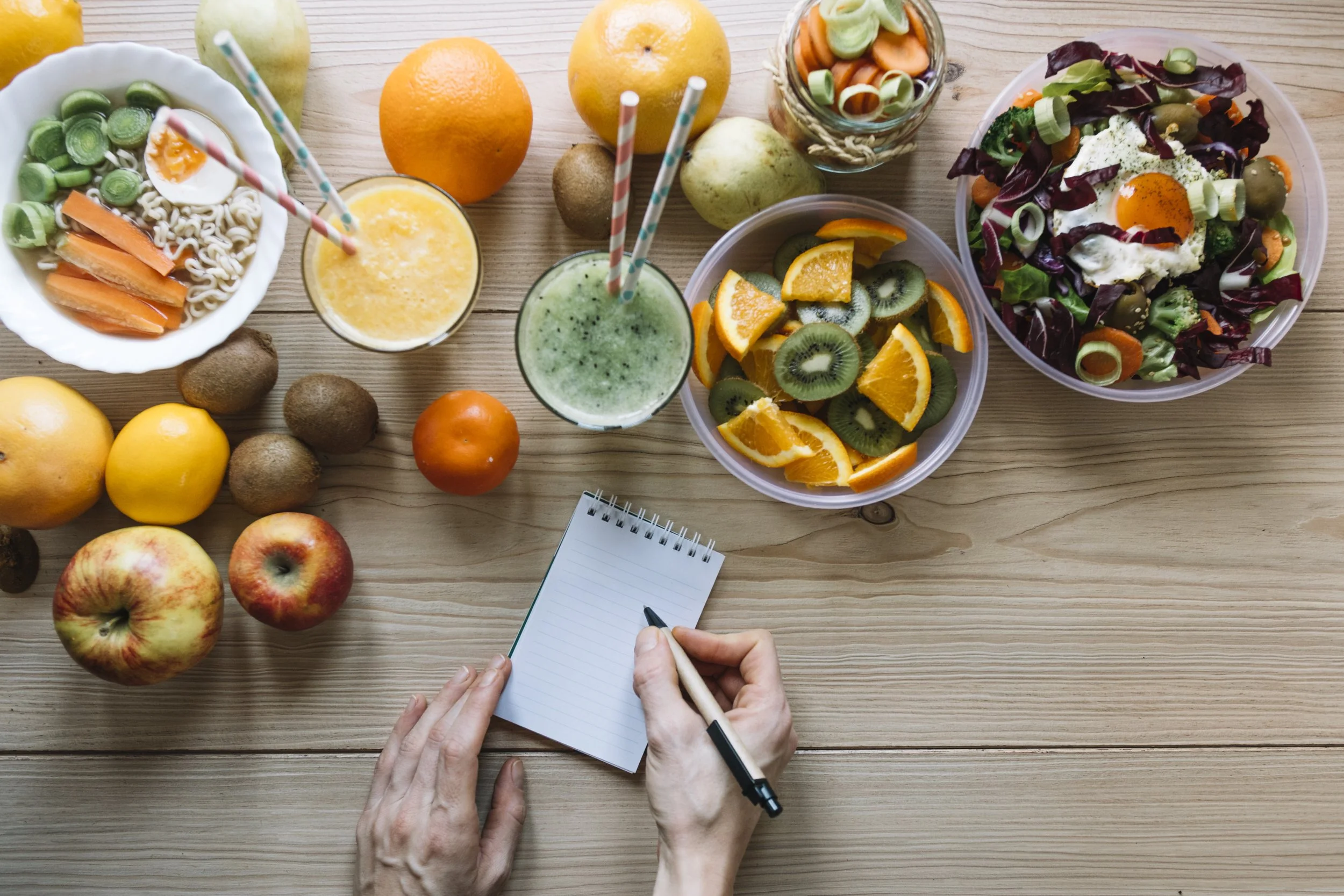 A variety of fresh fruits, salads, and drinks on a wooden table, with a person's hands writing in a small notepad.