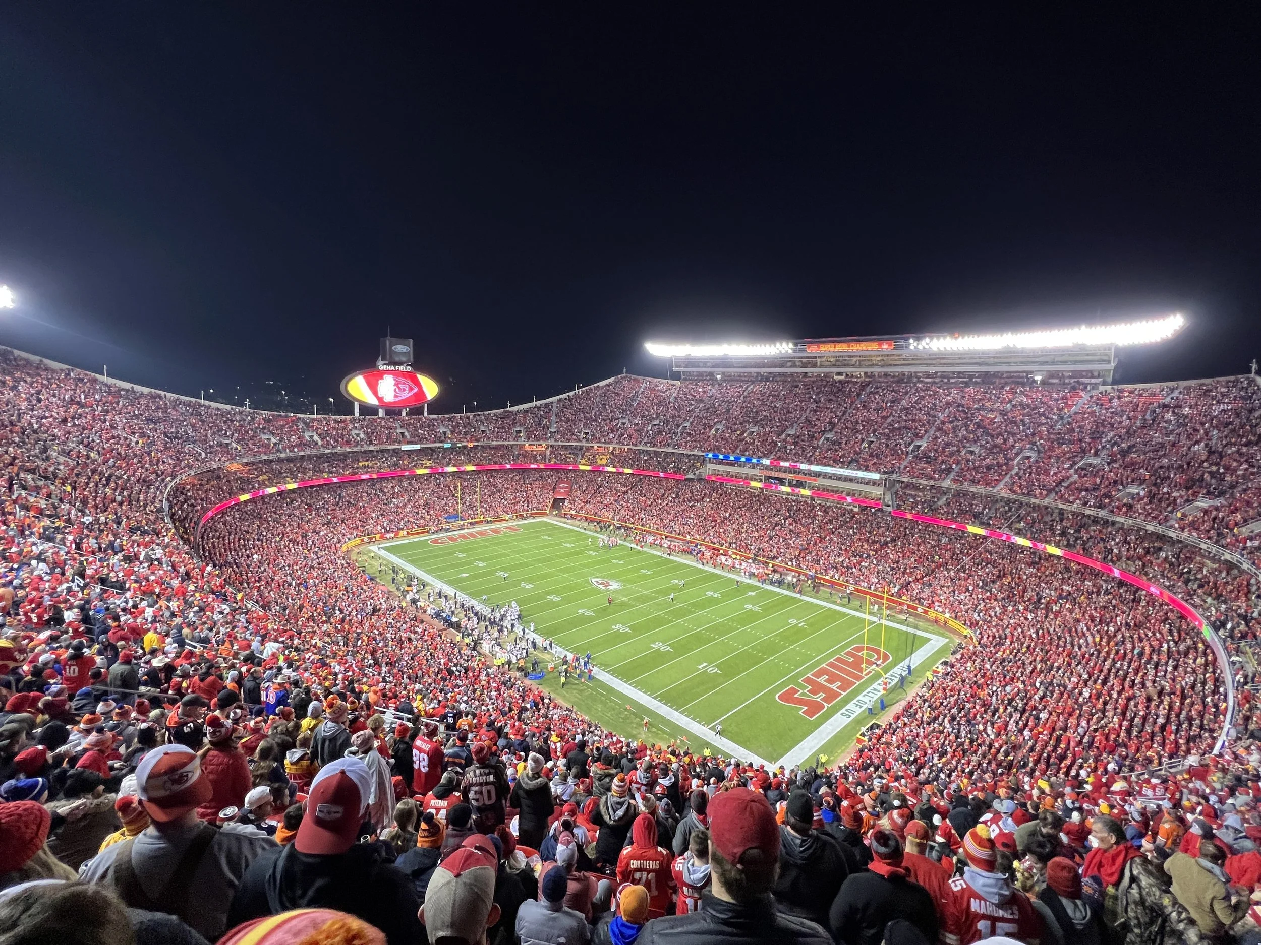 Nighttime view of a football stadium crowded with spectators, illuminated and filled with red-clad fans, with players on the field.
