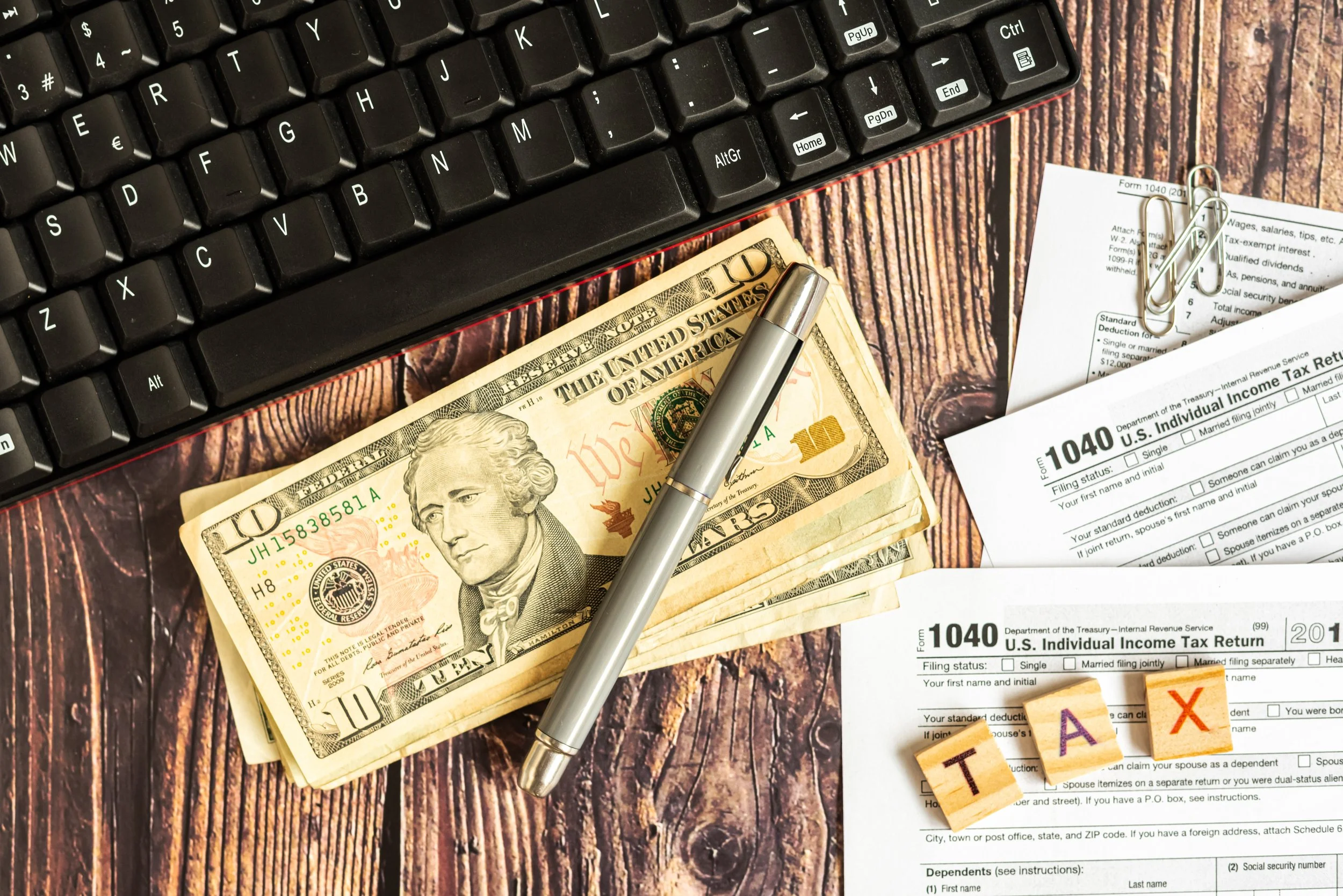 A desk with a black keyboard, a stack of ten dollar bills, a silver pen, tax forms, and wooden blocks spelling 'TAX' on a wooden surface.