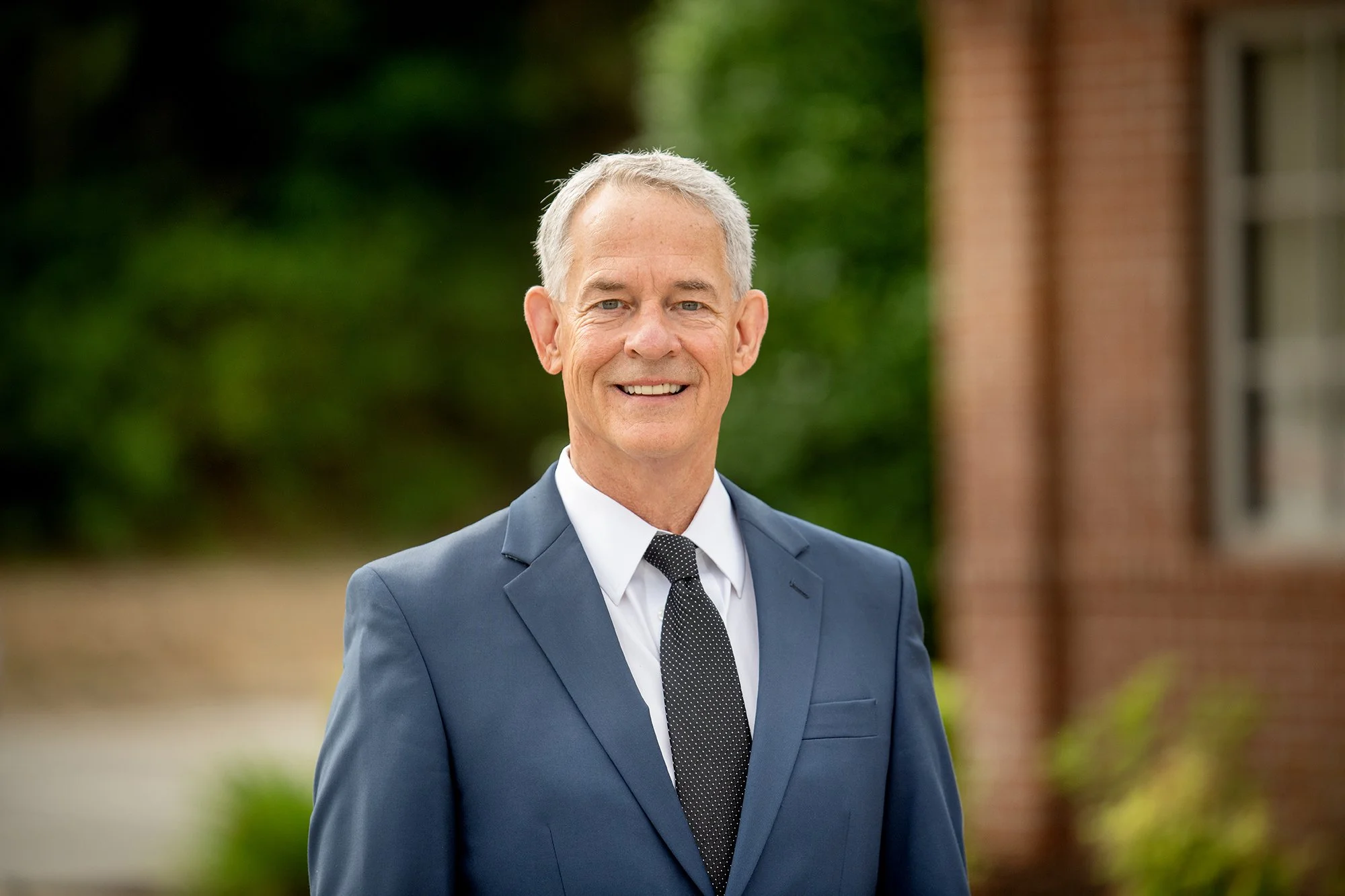 A smiling middle-aged man in a grey suit with a white shirt and a black dotted tie, standing outdoors near a brick building with greenery in the background.