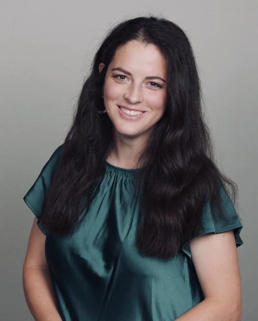 A woman with long dark hair smiling, wearing a teal blouse, standing against a plain light gray background.