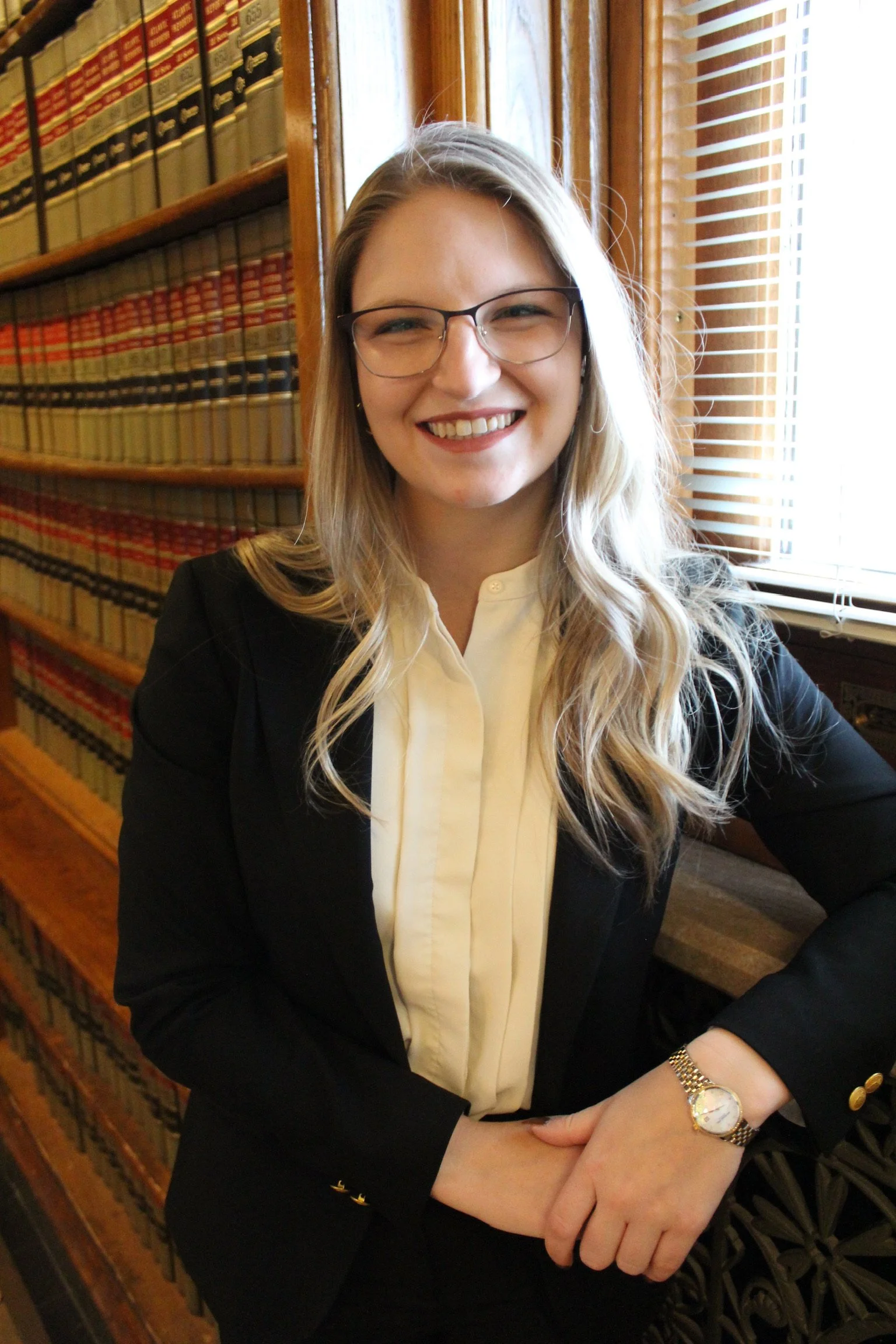 A woman with long blonde hair, glasses, and a white blouse under a black blazer, smiling in front of a wooden bookshelf filled with law books.
