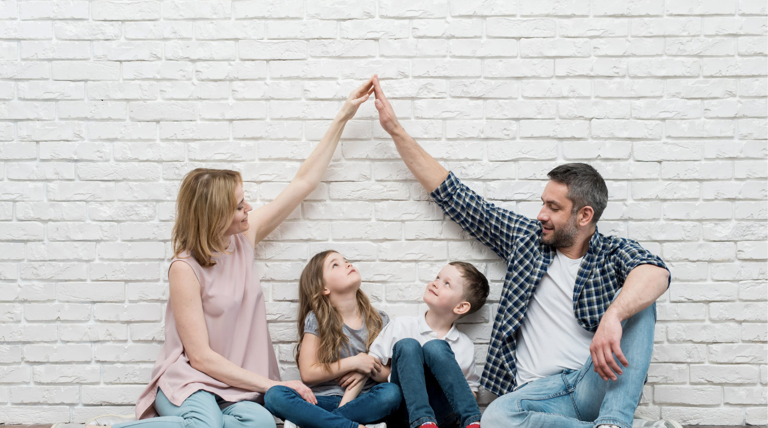 Família sentada no chão, fazendo um high five, com paredes de tijolos brancos ao fundo.