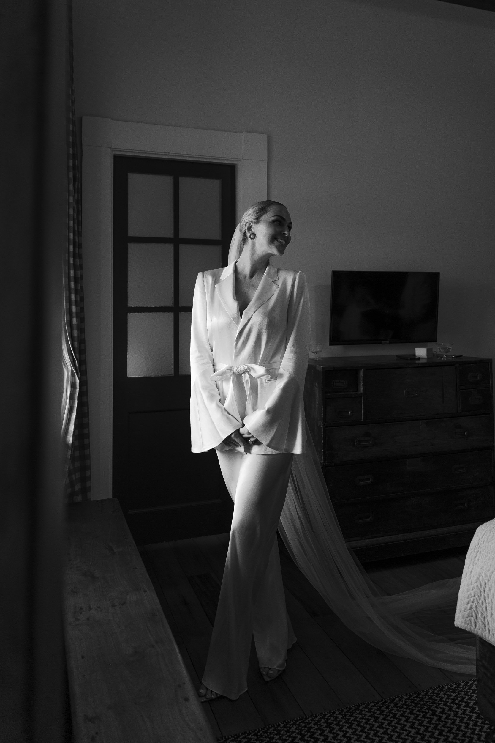 A woman in a light-colored suit with a satin lapel stands in a room, smiling and looking to her right. She has long hair and is wearing earrings. The room has a door with glass panes, a dark wooden dresser with a flat-screen TV on top, and appears to be part of a bedroom with a bed and a patterned rug.