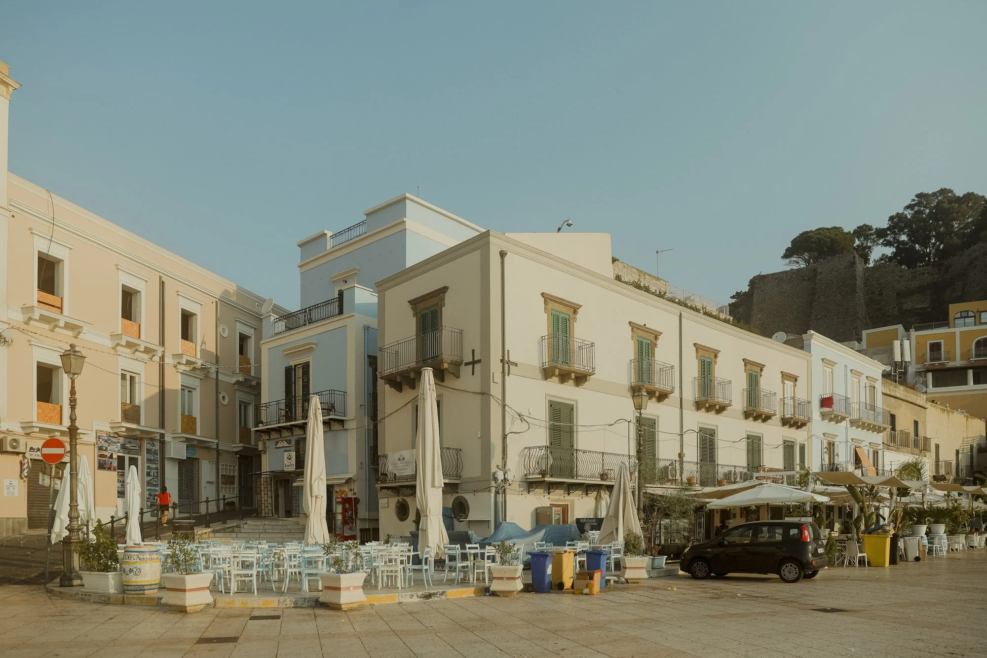 A sunny town square in Italy with pastel-colored buildings, outdoor seating with white chairs and umbrellas, and a black car parked in front.
