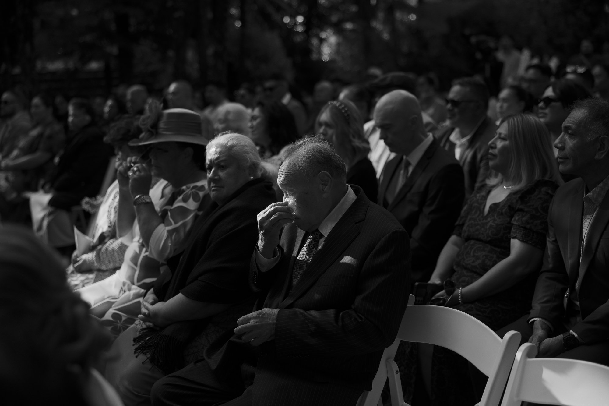 Black and white photo of a group of people seated outdoors, some crying or contemplative, attending a wedding in West Auckland.