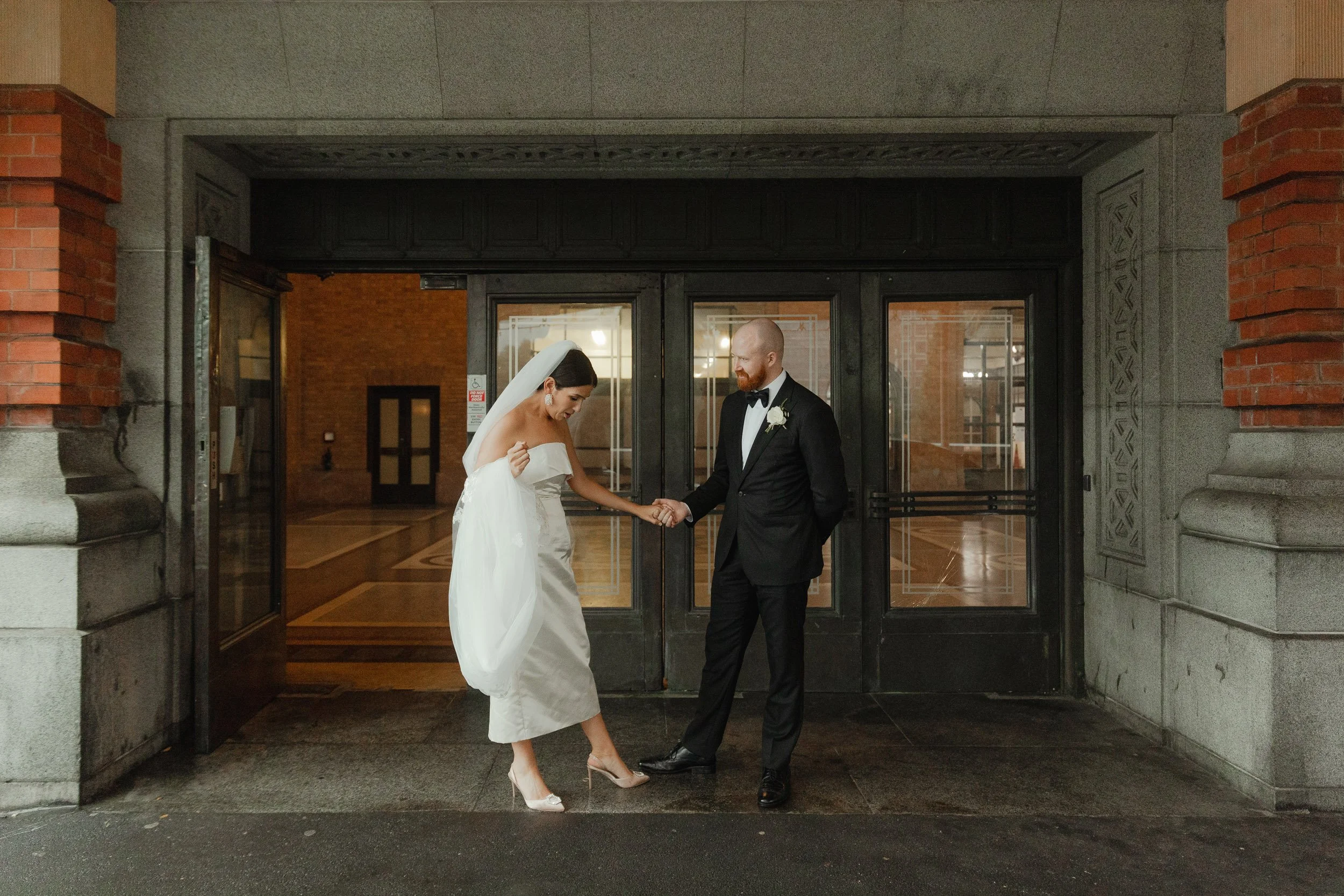 Bride and groom standing outside a building holding hands, with the bride lifting her dress slightly. The bride is wearing a white off-the-shoulder wedding gown and veil, and the groom is in a black tuxedo with a bow tie and boutonniere.