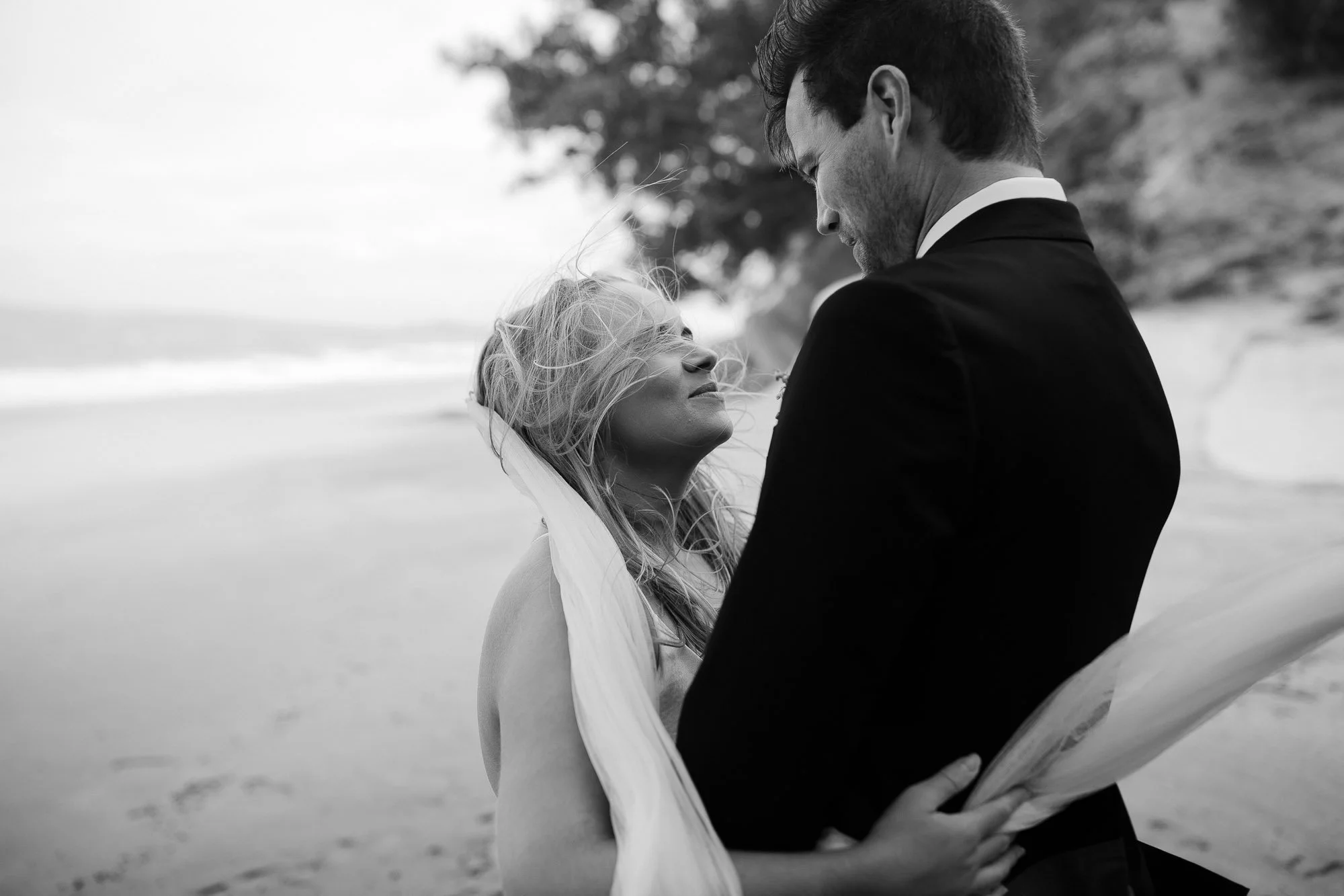 A black-and-white photo of a bride and groom on the beach, close together, sharing an intimate moment. The bride has her eyes closed and is smiling softly, while the groom looks down at her with a tender expression.
