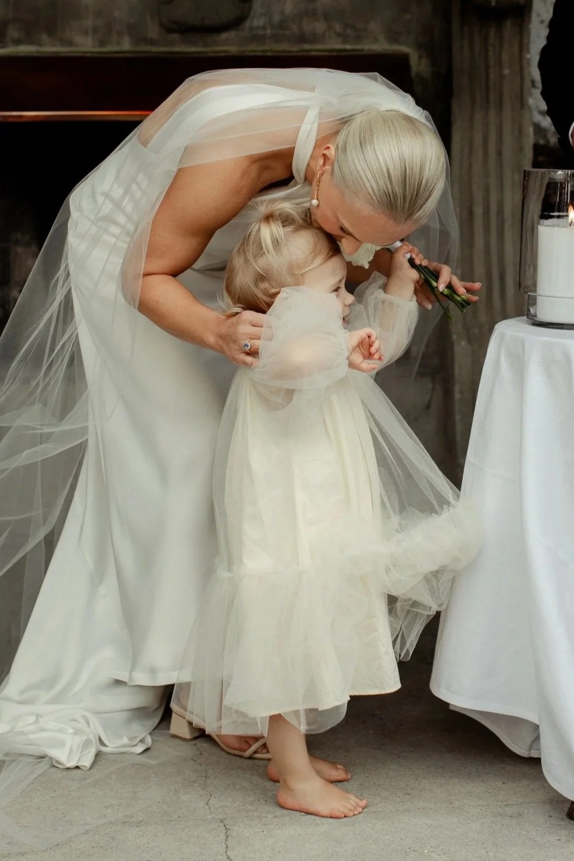 A bride, leaning over to kiss a young flower girl at a wedding ceremony. Both are dressed in white, with the girl wearing a tulle dress.