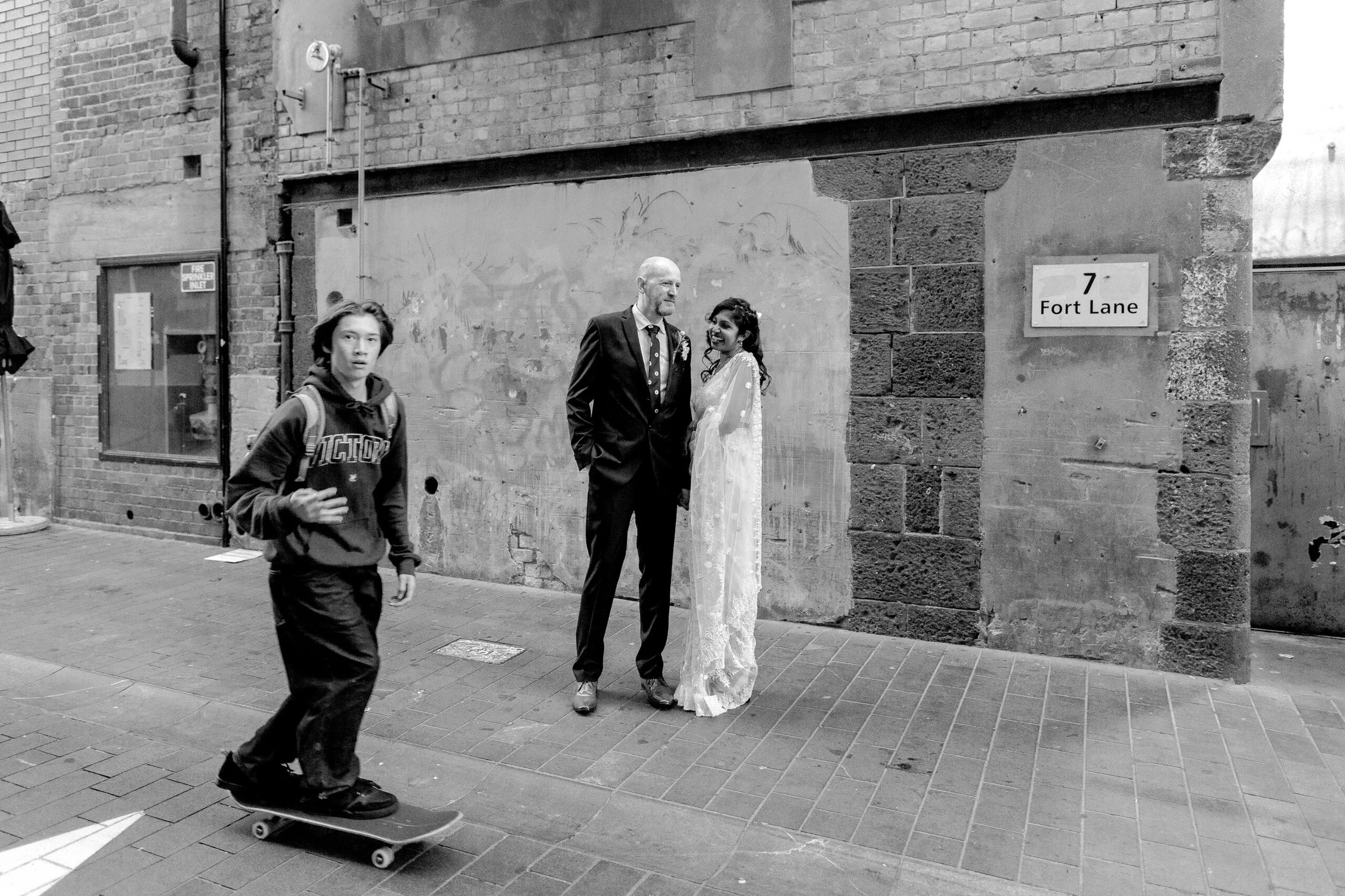 A skateboarder is skating past a couple standing and talking on an Auckland city street called Fort Lane. They are wearing a suit and a wedding dress after just getting married.