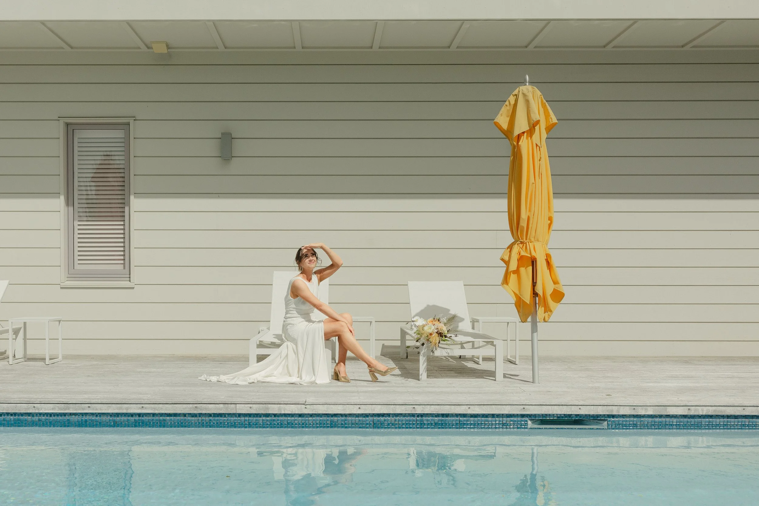 A woman in a white wedding dress sits on a poolside lounge chair, shielding her eyes from the sun, with a bouquet and a yellow umbrella nearby.