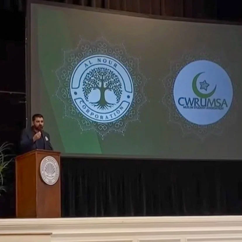 A man giving a speech at a podium in front of a large screen displaying two logos: one for Al Nour Corporation with a tree emblem, and another for CWRUMSA with a crescent moon and star emblem, during a conference or presentation.