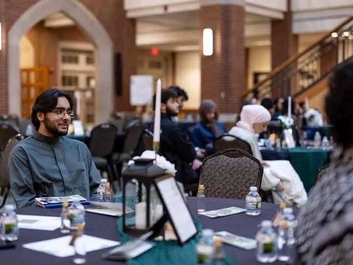 People sitting at tables in a conference or seminar room, with bottled water and documents on the tables.