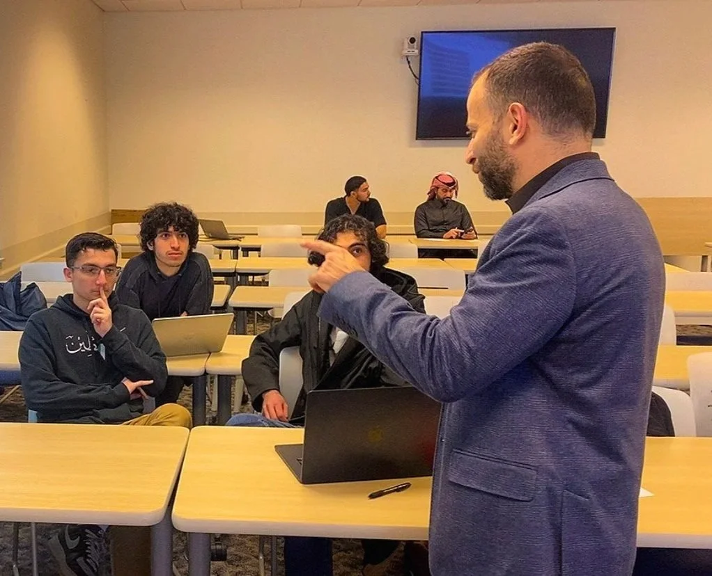 A man in a gray blazer is speaking to a classroom of students, gesturing with his finger. The students are seated at desks, some with laptops, listening attentively. The classroom has a large television screen on the wall.