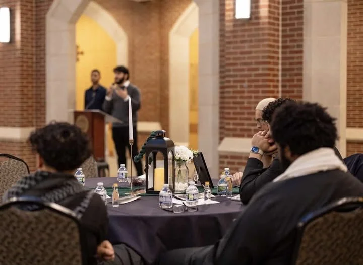 People seated around a round conference table with water bottles, glasses, and decorative items, listening to a speaker at a podium in a brick-walled room with arched doorways.