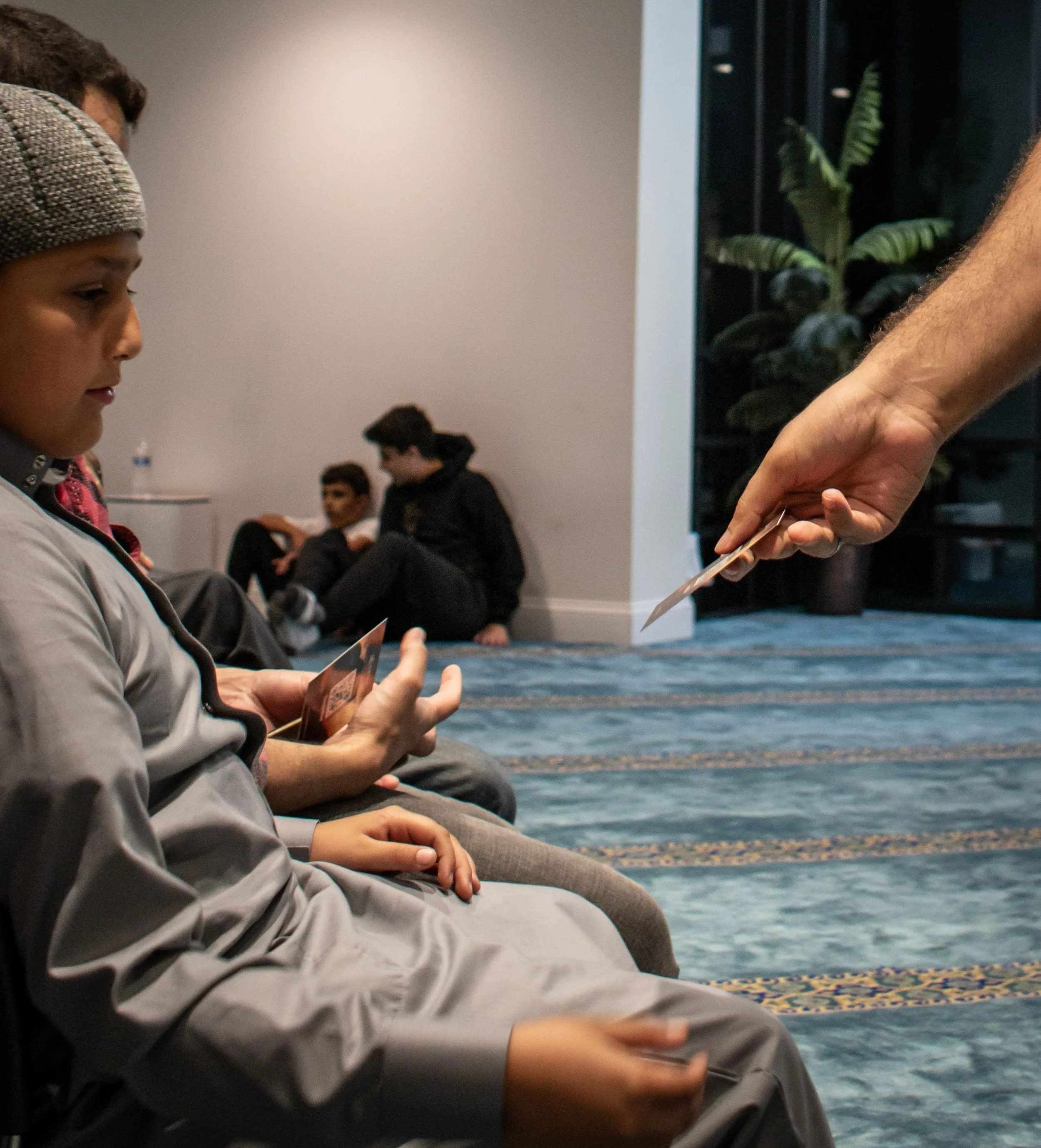 People sitting on the floor in a room, engaging in an activity involving passing cards or photographs to each other. The room has a patterned carpet, a plain wall, and a visible plant in the background.