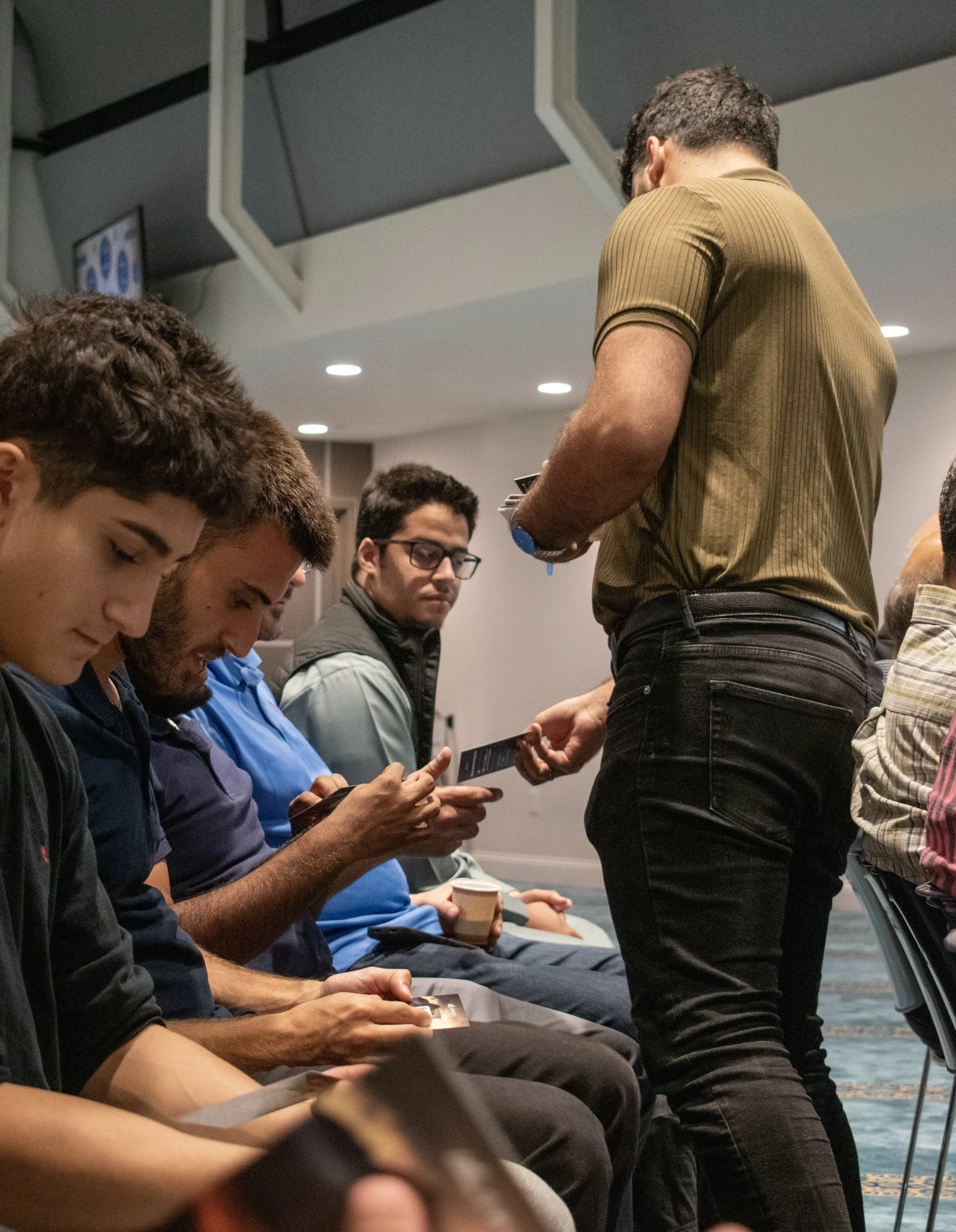 A group of young men sitting and looking at their phones or devices, while a woman stands in front of them holding a booklet or pamphlet.