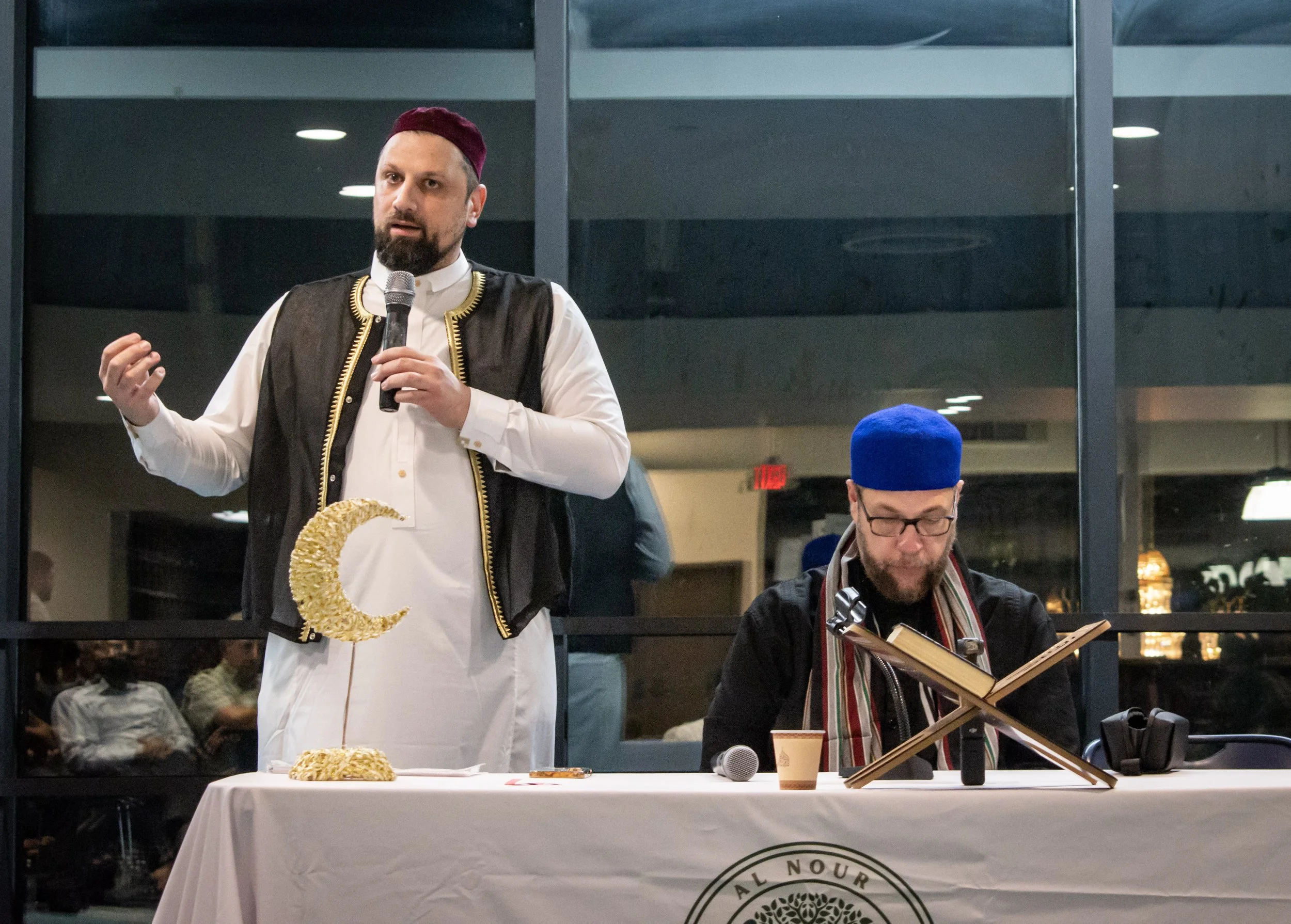 A man dressed in traditional Middle Eastern attire, holding a microphone, standing at a table with a decorative crescent moon and food items, with another man reading a book nearby in an indoor setting.