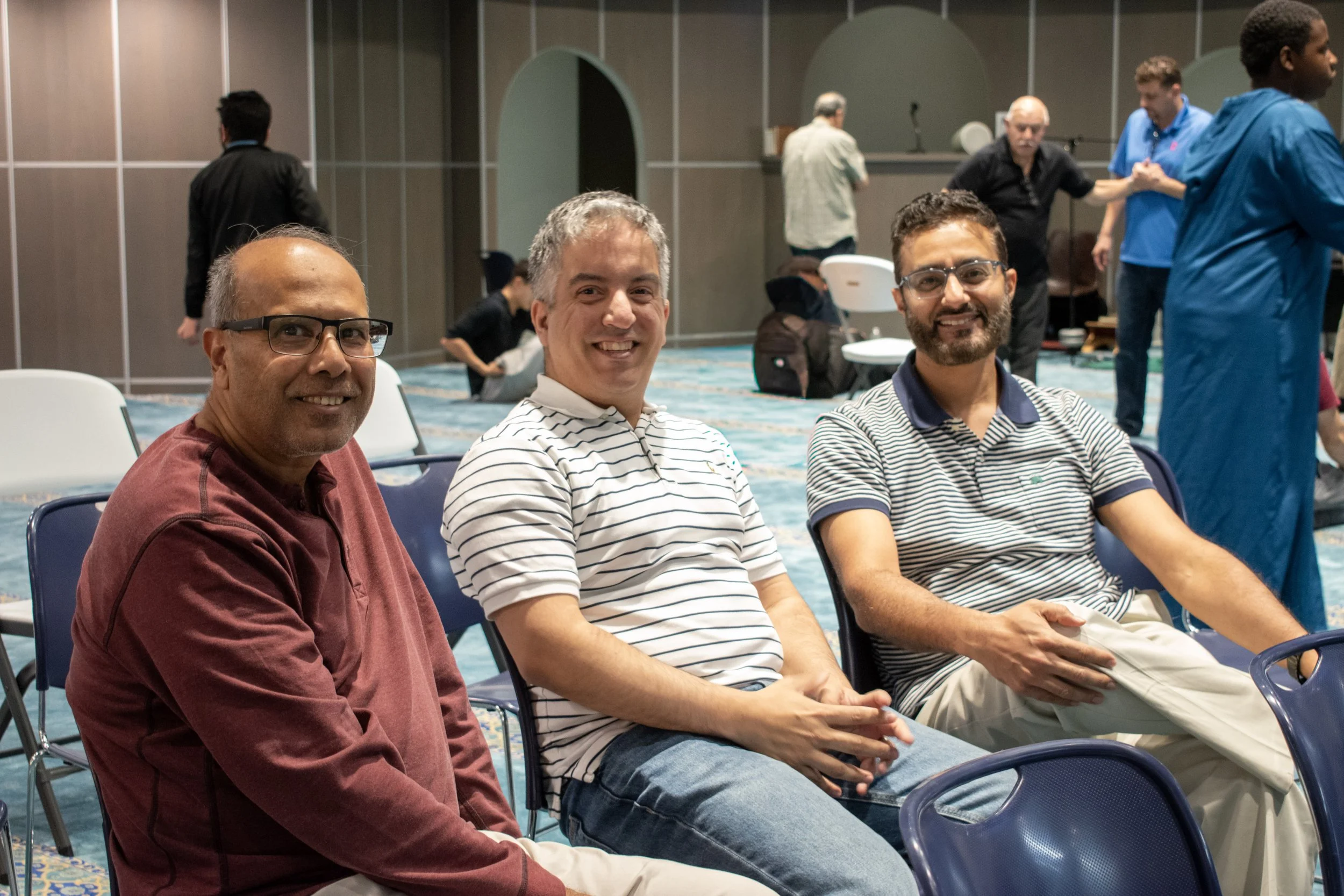Three men sitting and smiling at a conference or event, with people in the background engaged in conversations and activities.