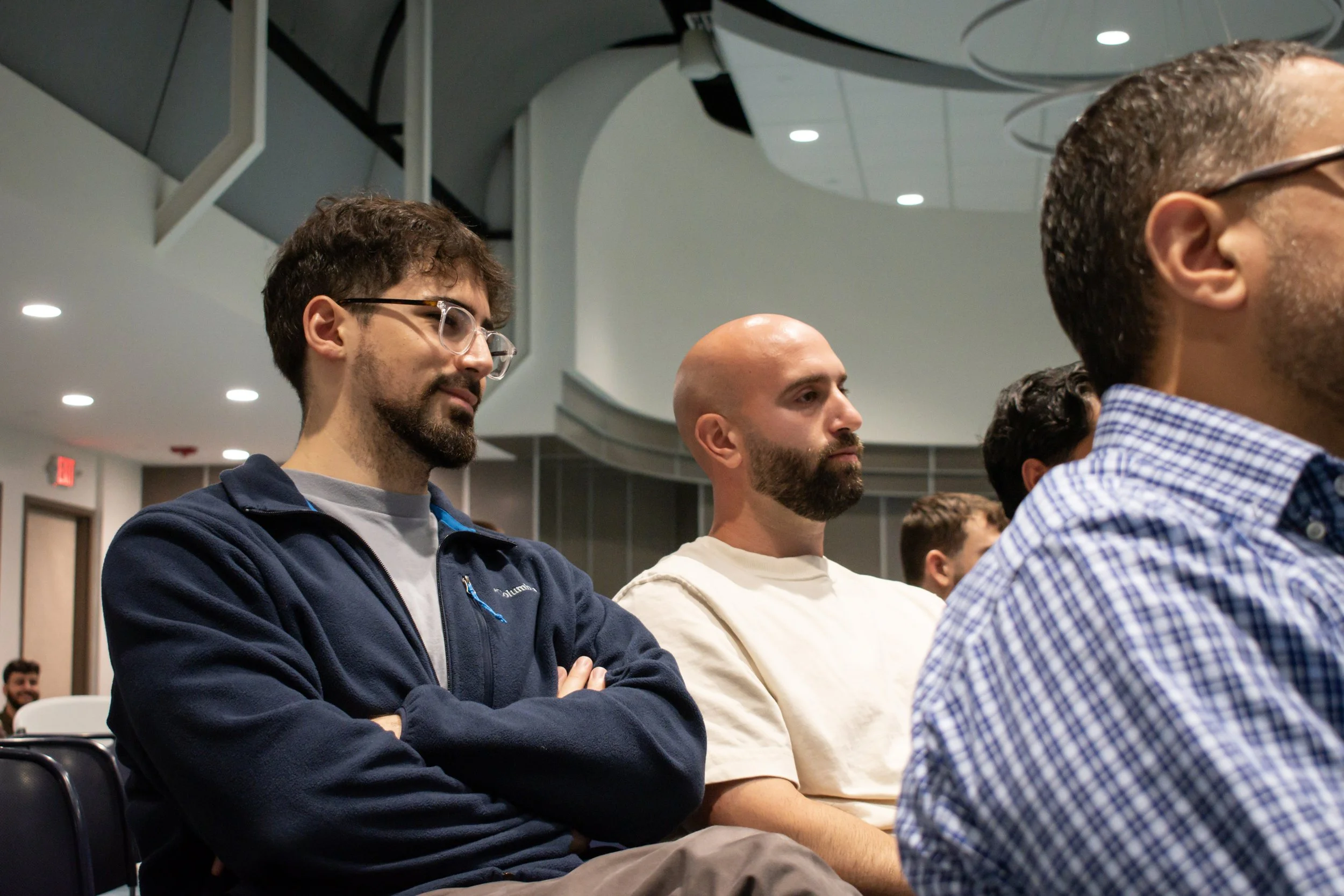 A group of men sitting in a lecture hall, attentively listening to a presentation. The men include a man with curly hair, glasses, and a beard wearing a dark hoodie, a bald man with a beard in a light shirt, and a man with short hair, glasses, and a checkered shirt. The background shows ceiling lights and a modern auditorium setting.