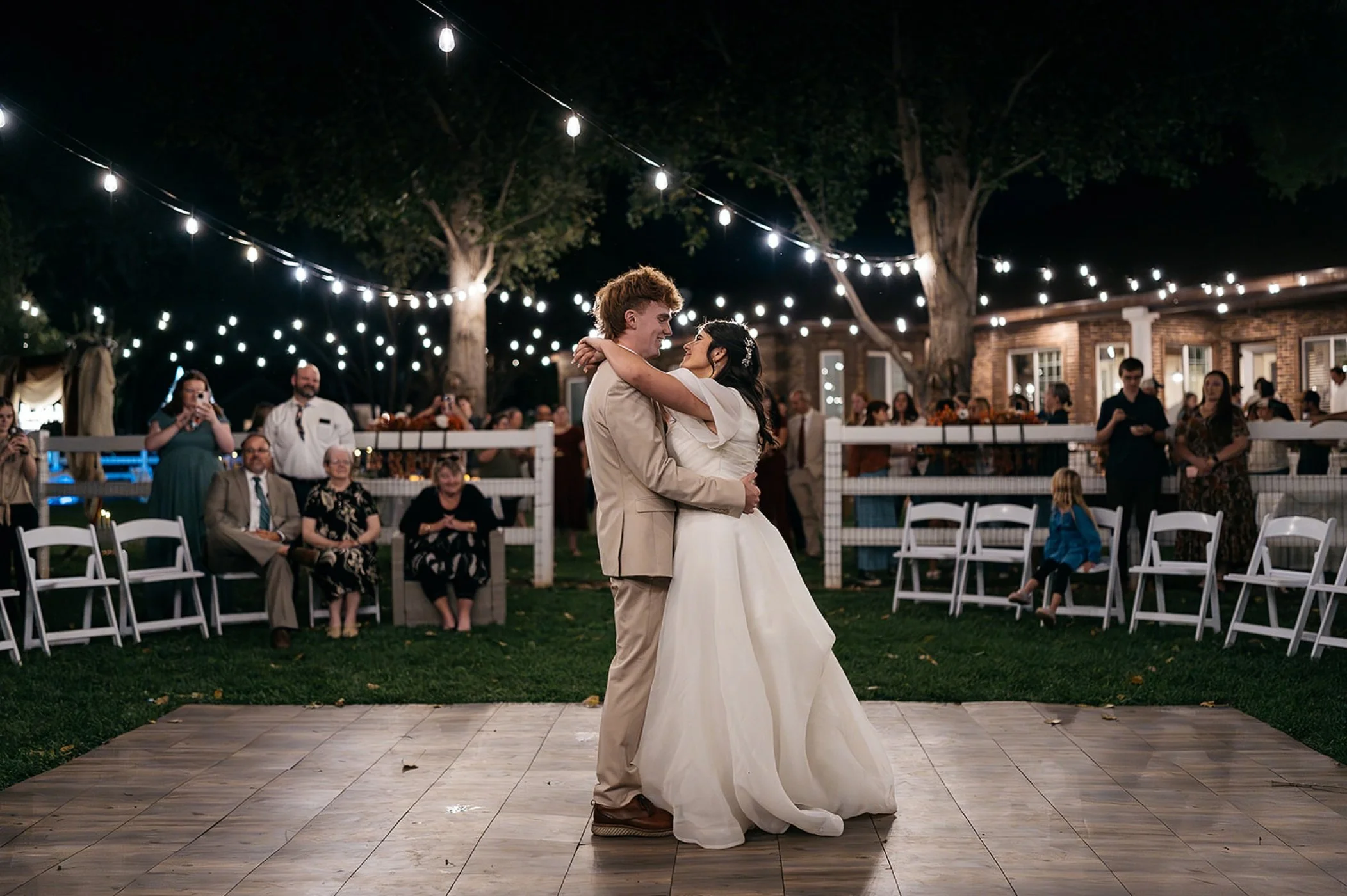 A bride and groom dancing at their outdoor wedding reception at night, under string lights, with guests sitting and standing around them. Dream Day Event Rentals, Vintage and Modern Decor and Floral Rentals, Freeport Illnois.