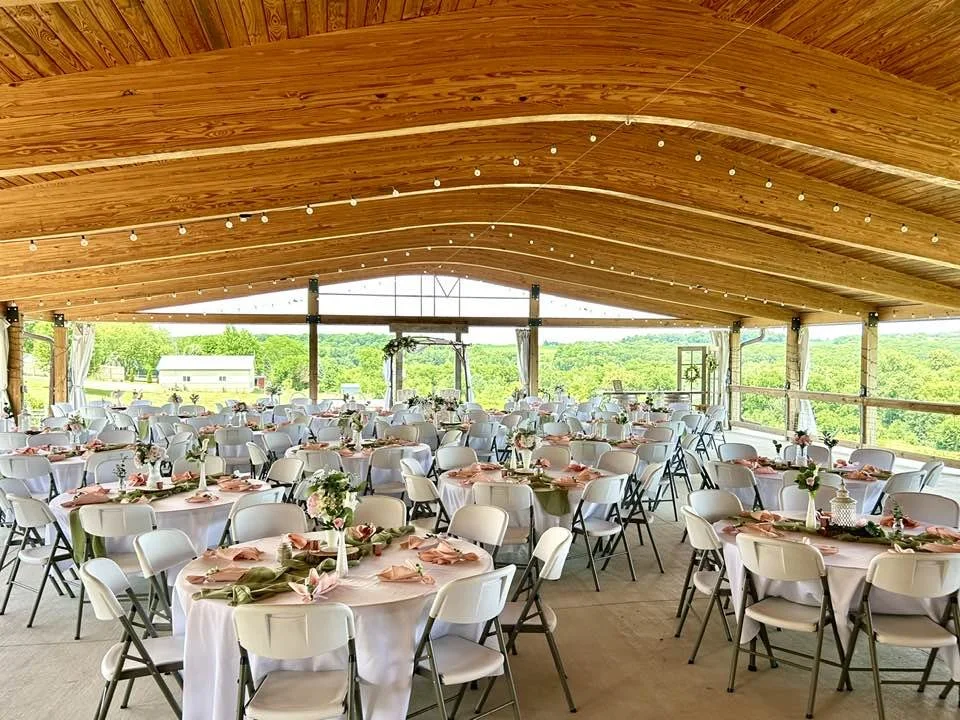Large rustic banquet hall with round tables covered in white tablecloths, decorated with pink and green napkins and flower arrangements. The hall has a wooden arched ceiling with string lights and large open windows showing a scenic view of green tre