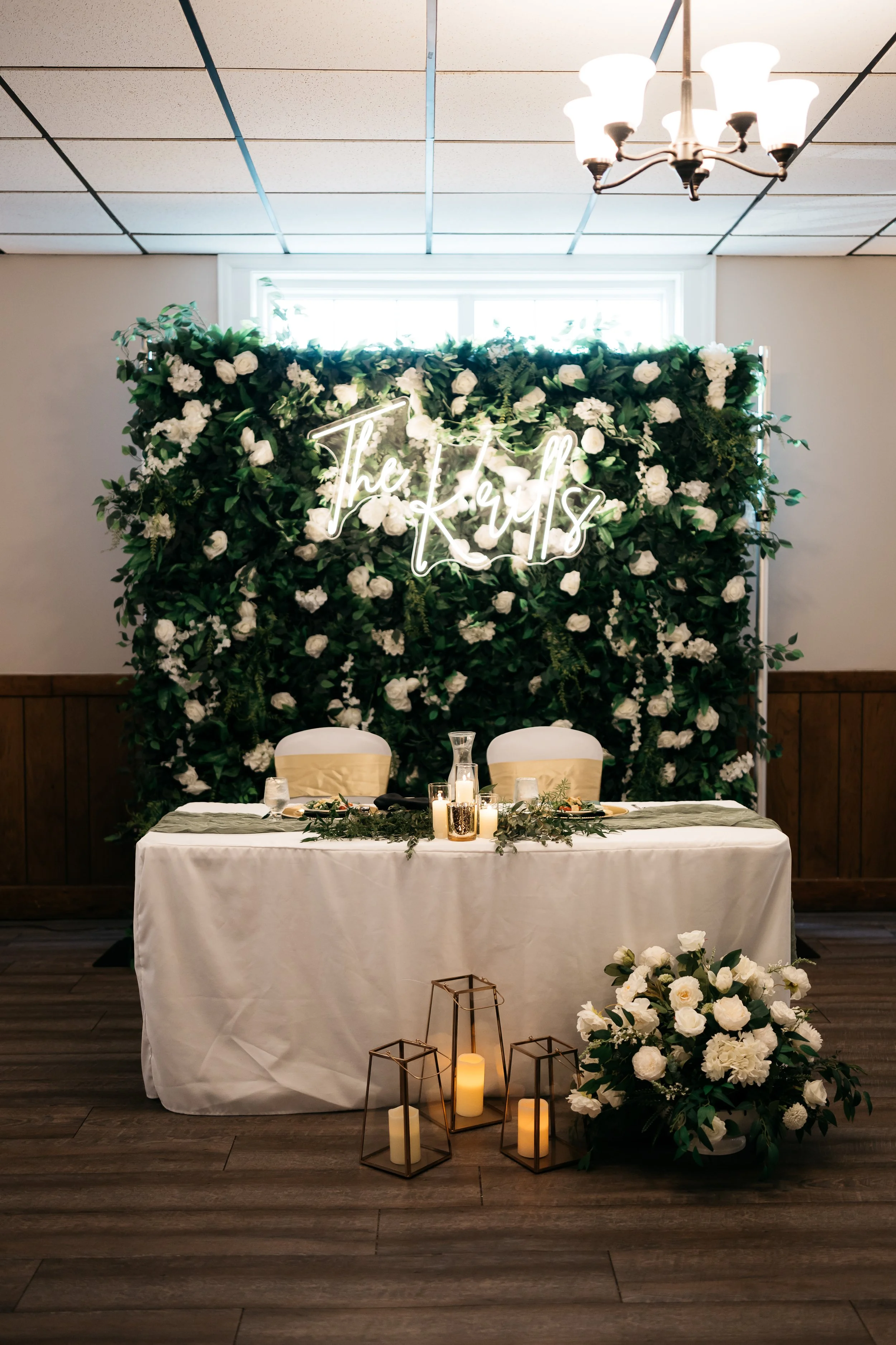 Wedding reception setup with a table, floral arrangements, candles, and a illuminated sign that reads 'The Keys' against a green and white floral backdrop. Dream Day Event Rentals, Vintage and Modern Decor and Floral Rentals, Freeport Illnois.