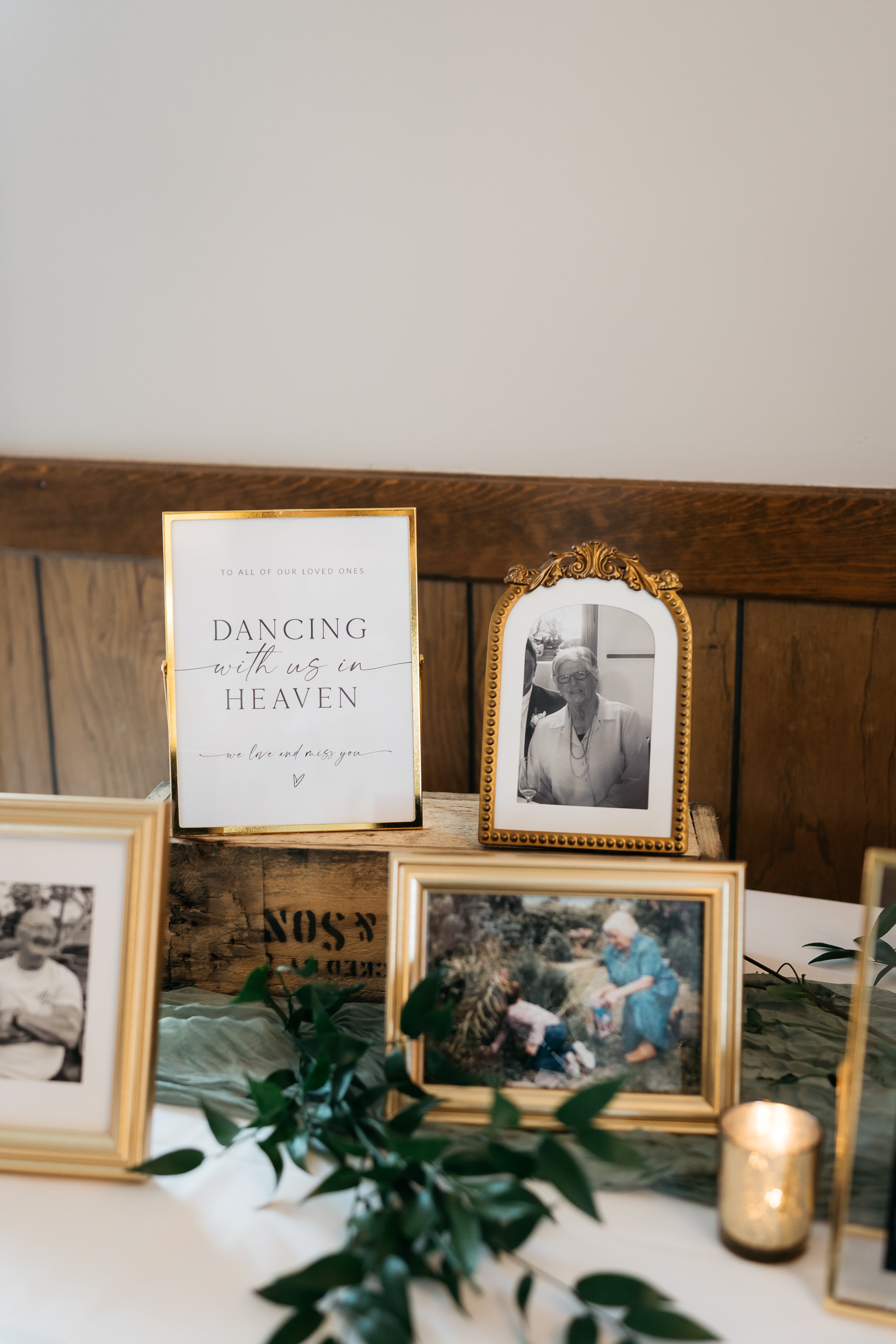 A memorial table with framed photos and a sign, including a black-and-white photo of an elderly woman, two vintage-style color photos, and a candle, decorated with green foliage. Dream Day Event Rentals, Vintage and Modern Decor and Floral Rentals.