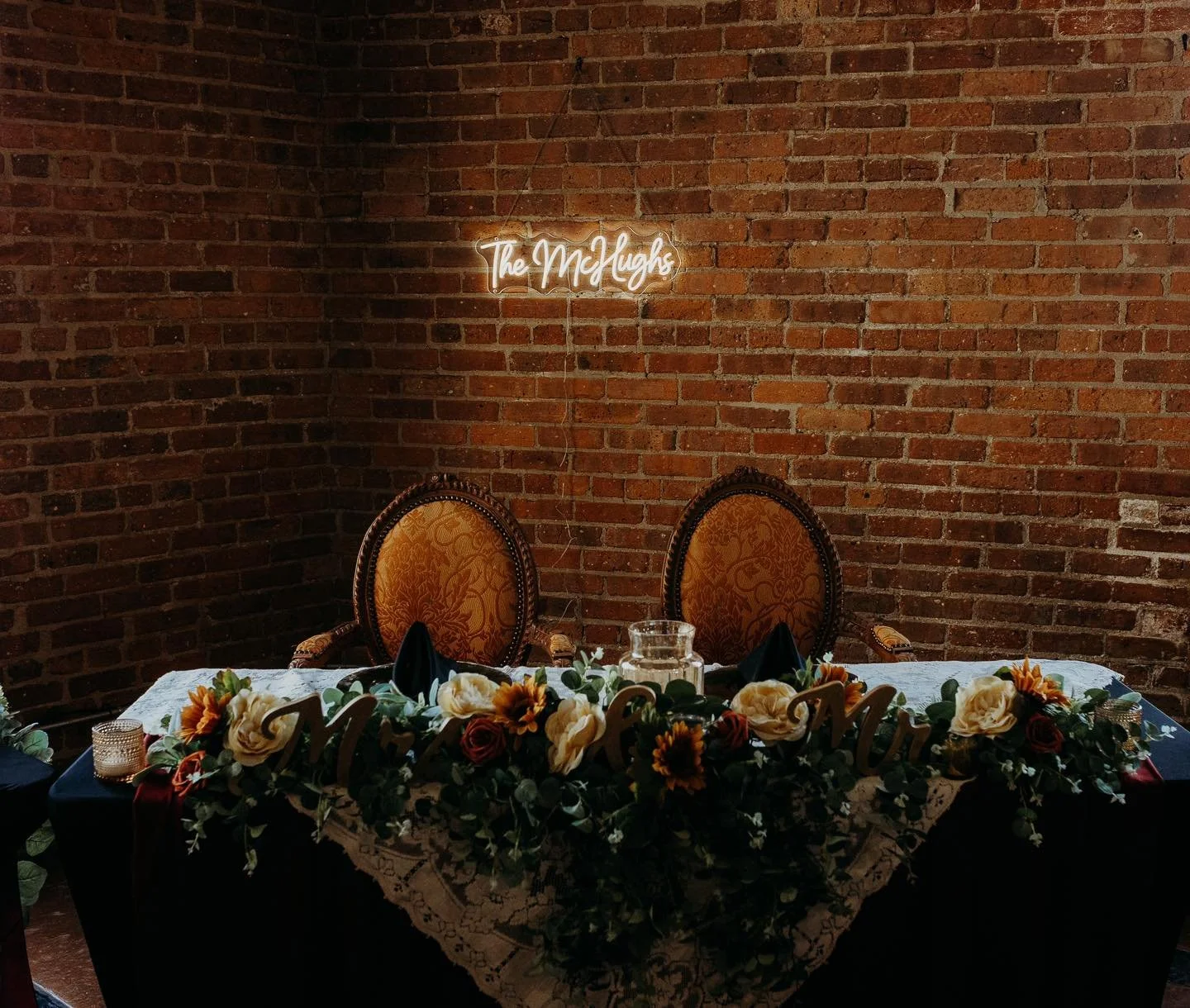 A decorated table with floral arrangements, a lace tablecloth, and Halloween-themed signs, set against an exposed brick wall. Dream Day Event Rentals, Vintage and Modern Decor and Floral Rentals