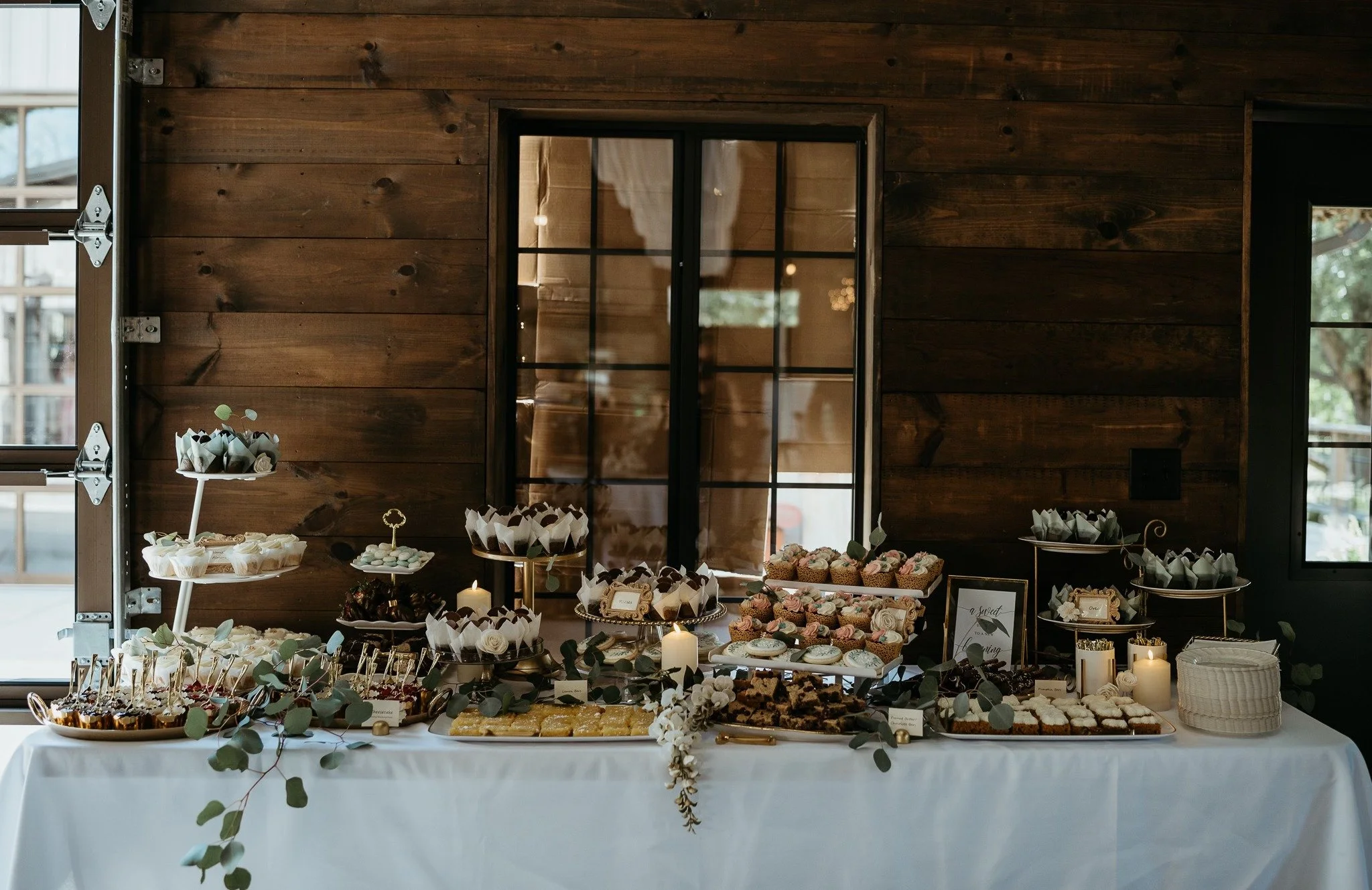 Dessert table decorated with greenery, candles, and assorted sweets including cupcakes, cookies, and chocolates, set against a dark wooden wall and a window. Dream Day Event Rentals, Vintage and Modern Decor and Floral Rentals, Freeport Illnois.