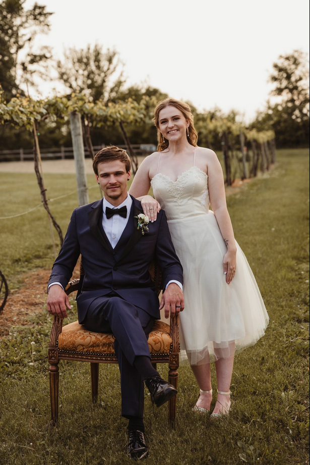 A bride and groom outdoors celebrating, groom seated in a vintage chair wearing a dark tuxedo, bride standing beside him with her hand on his shoulder, wearing a white dress, in a garden with greenery and trees.