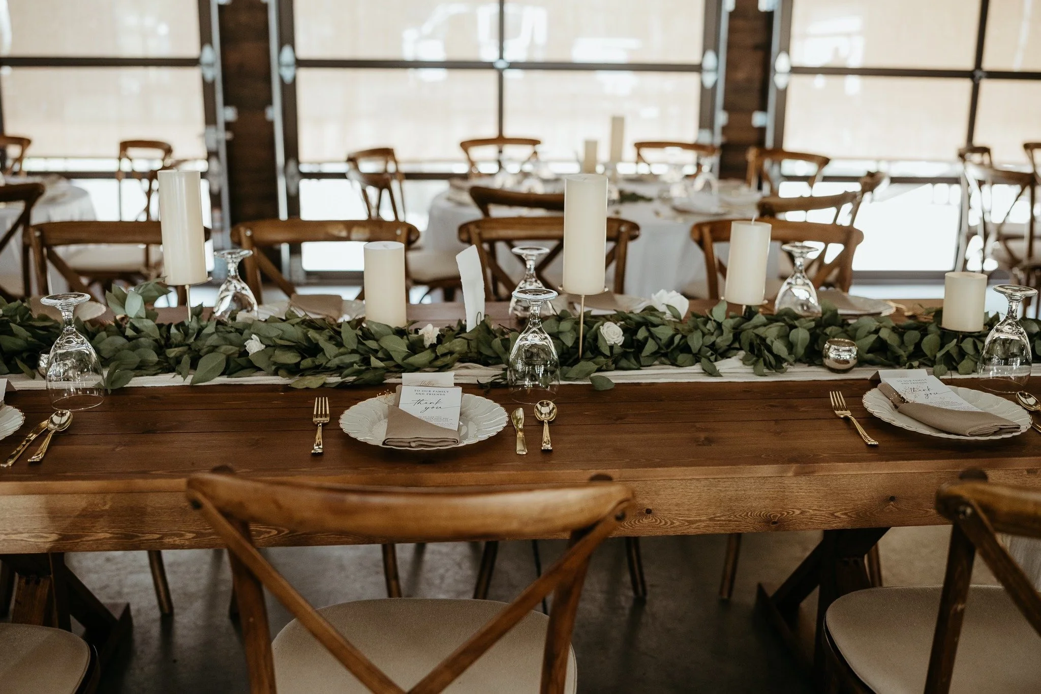 Wedding reception table decorated with candles, greenery, place settings, and chairs in a rustic-style venue with large windows. Dream Day Event Rentals, Vintage and Modern Decor and Floral Rentals, Freeport Illnois.