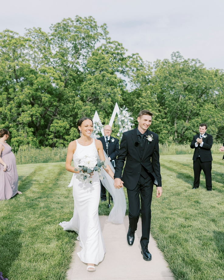 A wedding couple holding hands and walking down an outdoor aisle, with smiling guests clapping in the background, lush green trees, and a cloudy sky. Dream Day Event Rentals, Vintage and Modern Decor and Floral Rentals, Freeport Illnois.
