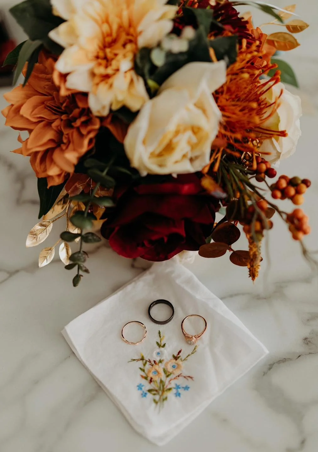 A bouquet of orange, white, and deep red flowers, with some gold leaves, placed on a marble surface. In front of the bouquet is a white embroidered cloth with a floral design, holding three rings, including two gold and one black.
