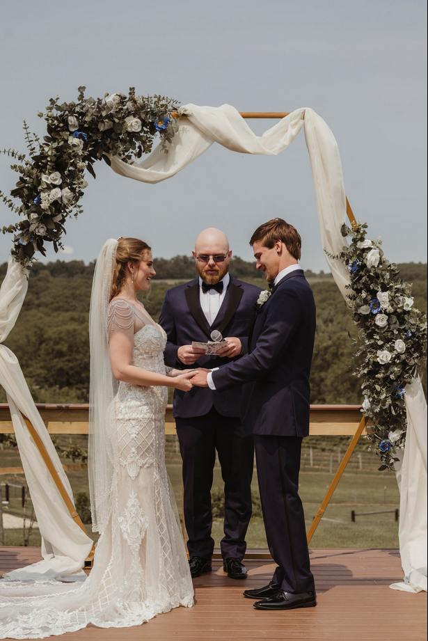 A bride and groom during their outdoor wedding ceremony, holding hands and smiling at each other, with an officiant reading vows under a decorated arch with white and blue flowers on a wooden platform.