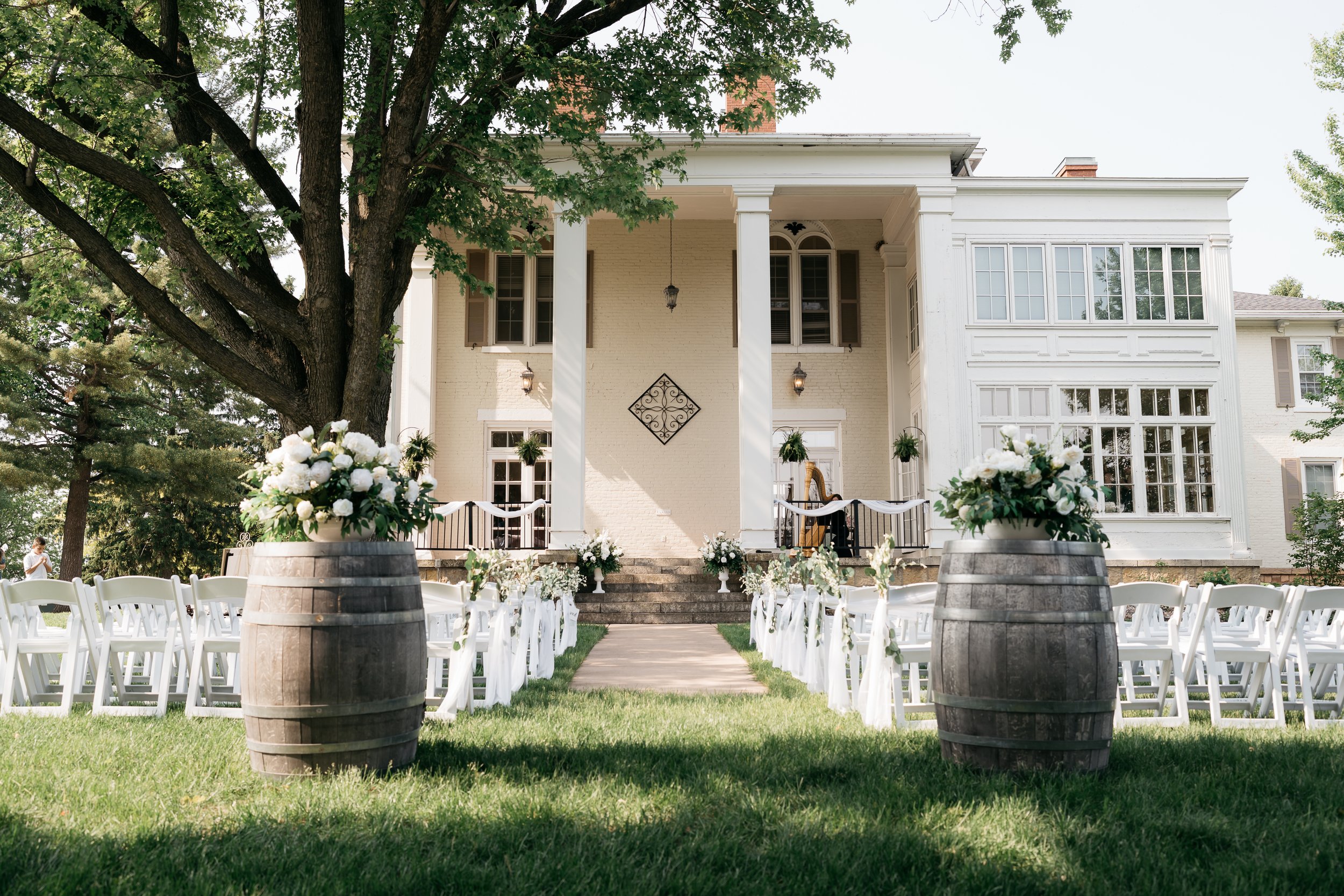 Outdoor wedding setup in front of a large white house with green trees, featuring white chairs, floral arrangements, and barrels with flowers, suggesting a wedding ceremony.