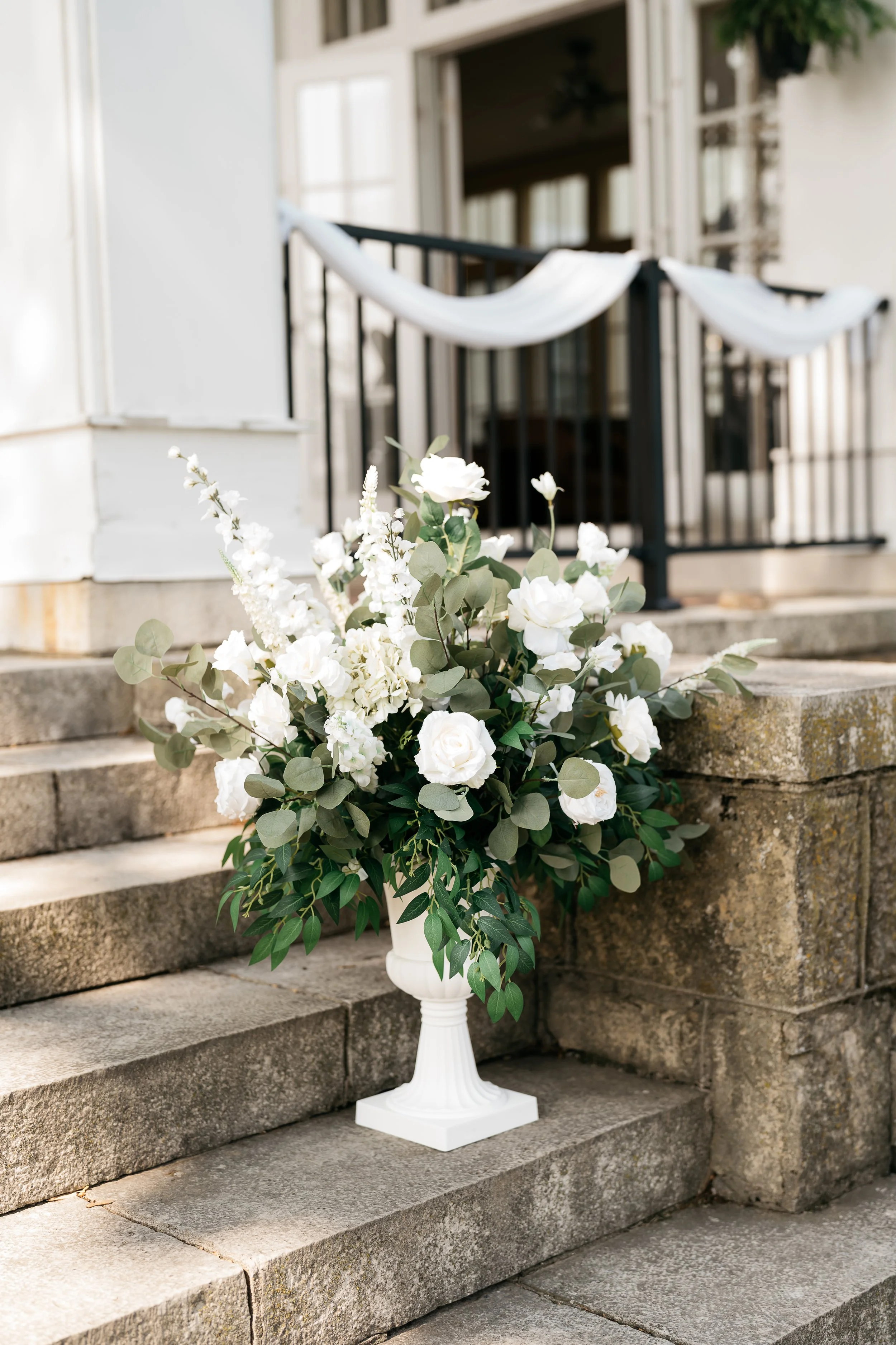 White flower arrangement in a decorative urn on stone steps outside a house with a porch. Dream Day Event Rentals, Vintage and Modern Decor and Floral Rentals, Freeport Illnois, and Madison Wisconsin