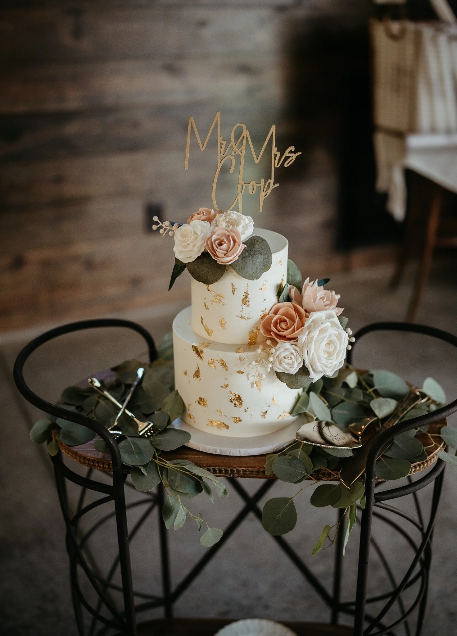 Two-tier wedding cake decorated with white and blush roses, greenery, and gold leaf accents, topped with a 'Mr & Mrs' cake topper, on a black wire cart decorated with eucalyptus. Dream Day Event Rentals, Vintage and Modern Decor and Floral Rentals.