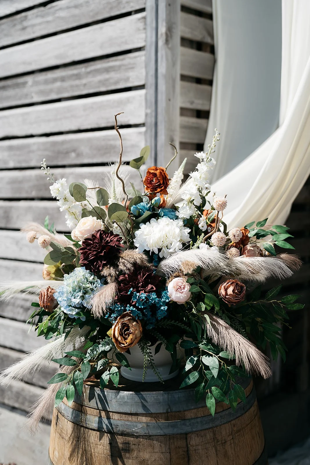 A bouquet of mixed flowers with white, brown, blue, and pink blooms, accented with feathers and green leaves, placed on a wooden barrel. Dream Day Event Rentals, Vintage and Modern Decor and Floral Rentals, Freeport, Illinois
