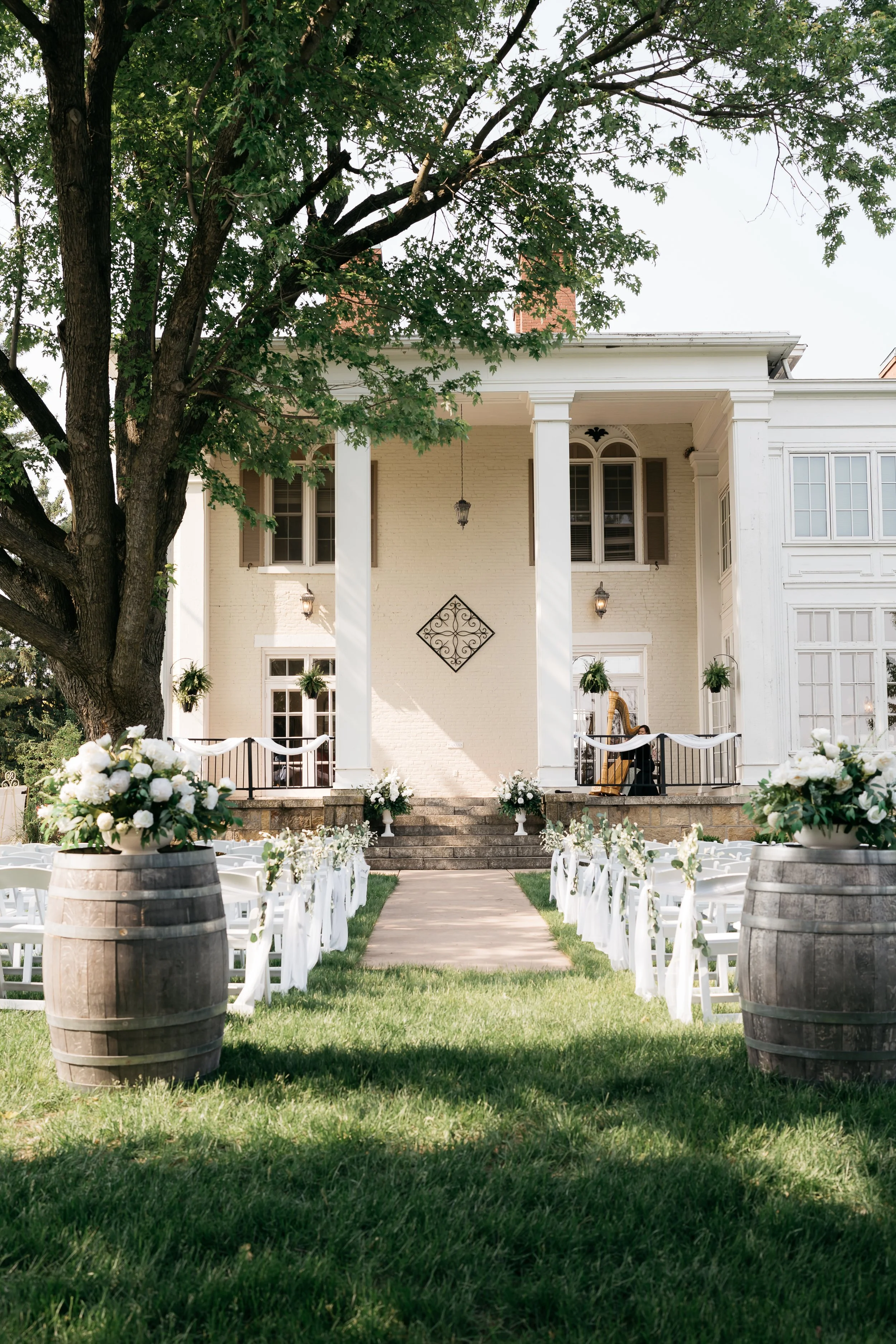 Outdoor wedding setup with white chairs decorated with white ribbons and flowers, wooden barrels with floral arrangements, stone steps leading up to a white house with large columns. Dream Day Event Rentals, Vintage and Modern Decor and Floral Rental