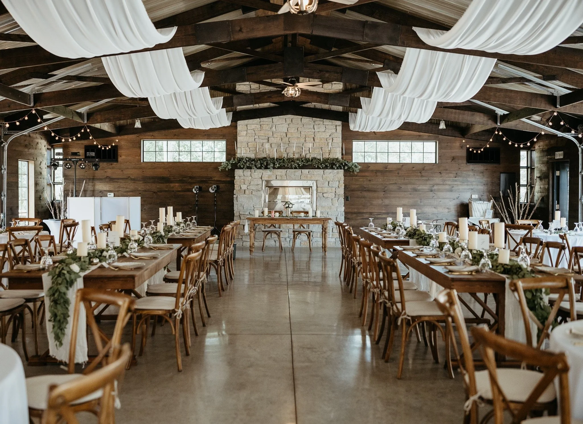 Elegant wedding reception setup in a rustic barn with long wooden tables decorated with white candles, candles, greenery, and white tableware. Dream Day Event Rentals, Vintage and Modern Decor and Floral Rentals, Freeport Il
