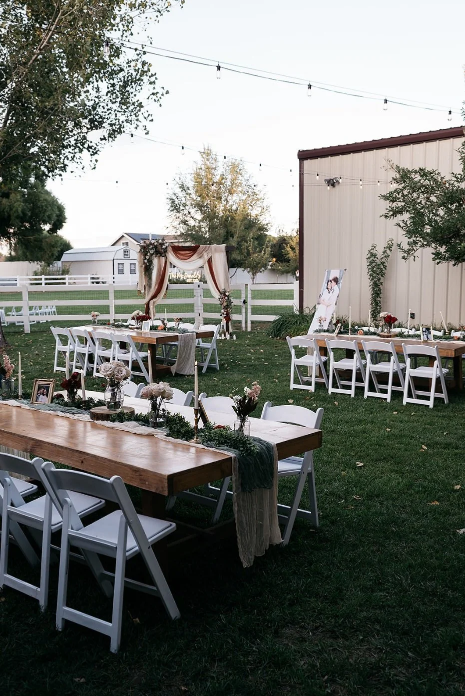 Outdoor wedding reception setup with tables decorated with flowers, a backdrop with drapes, and string lights overhead on a grass lawn. Dream Day Event Rentals, Vintage and Modern Decor and Floral Rentals, Freeport Illnois.
