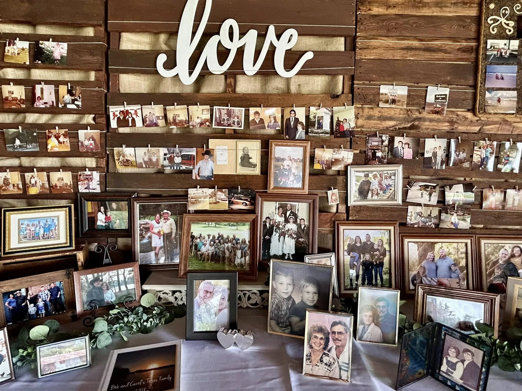 Display of framed photos and photo prints on a table and hanging on a wood-paneled wall, with a large decorative 'love' sign at the top. Dream Day Event Rentals, Vintage and Modern Decor and Floral Rentals, Freeport Illnois.