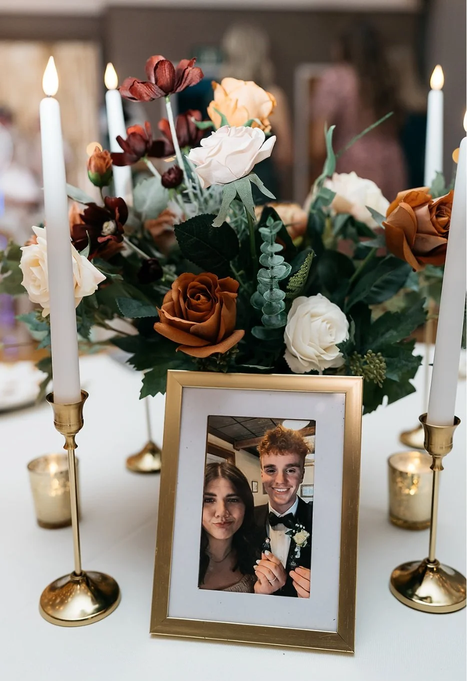 A table centerpiece with a floral arrangement of white, cream, orange, and dark red roses, surrounded by lit white candles in gold holders, and a framed photo of a smiling couple dressed formally, likely at a wedding or special event.
