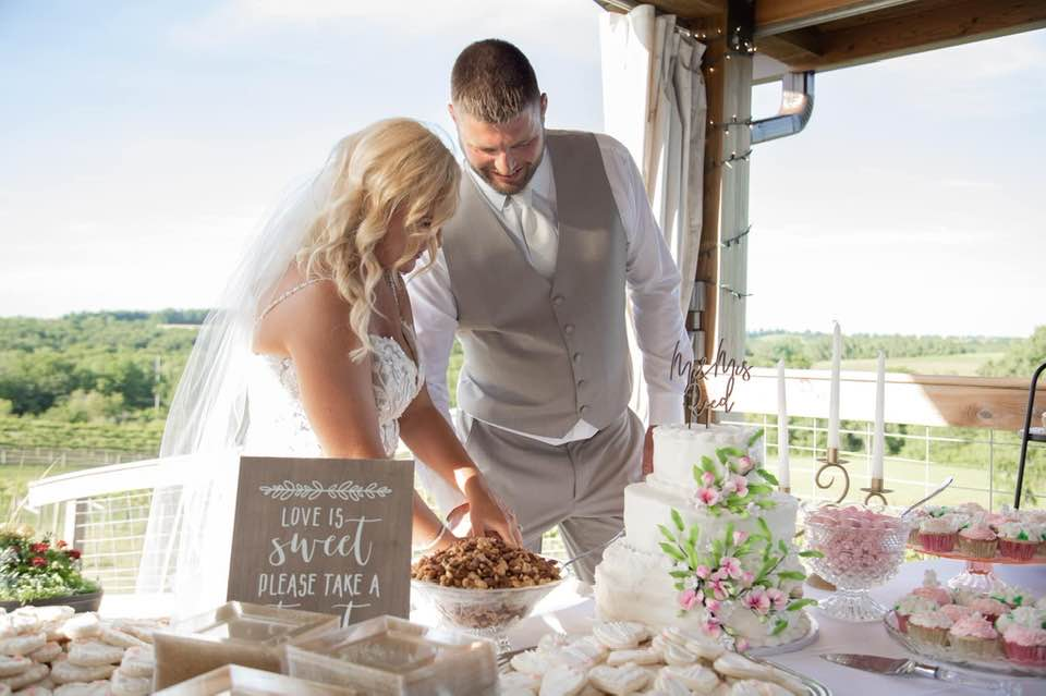 A bride and groom cutting their wedding cake outdoors on a sunny day with greenery in the background. Dream Day Event Rentals, Vintage and Modern Decor and Floral Rentals, Freeport Illnois.
