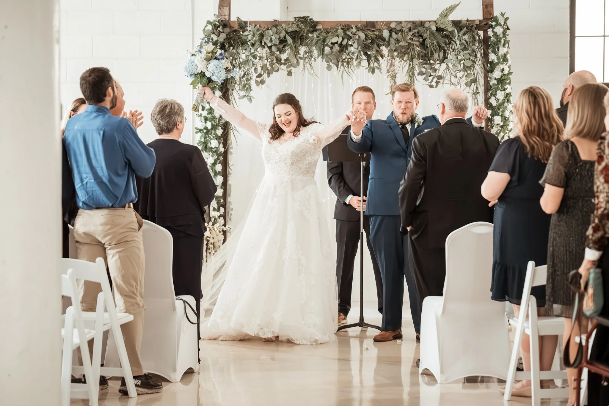 A bride and groom celebrating their wedding with raised hands, surrounded by family and friends in an indoor venue decorated with flowers and greenery. Dream Day Event Rentals, Vintage and Modern Decor and Floral Rentals, Freeport Illnois.
