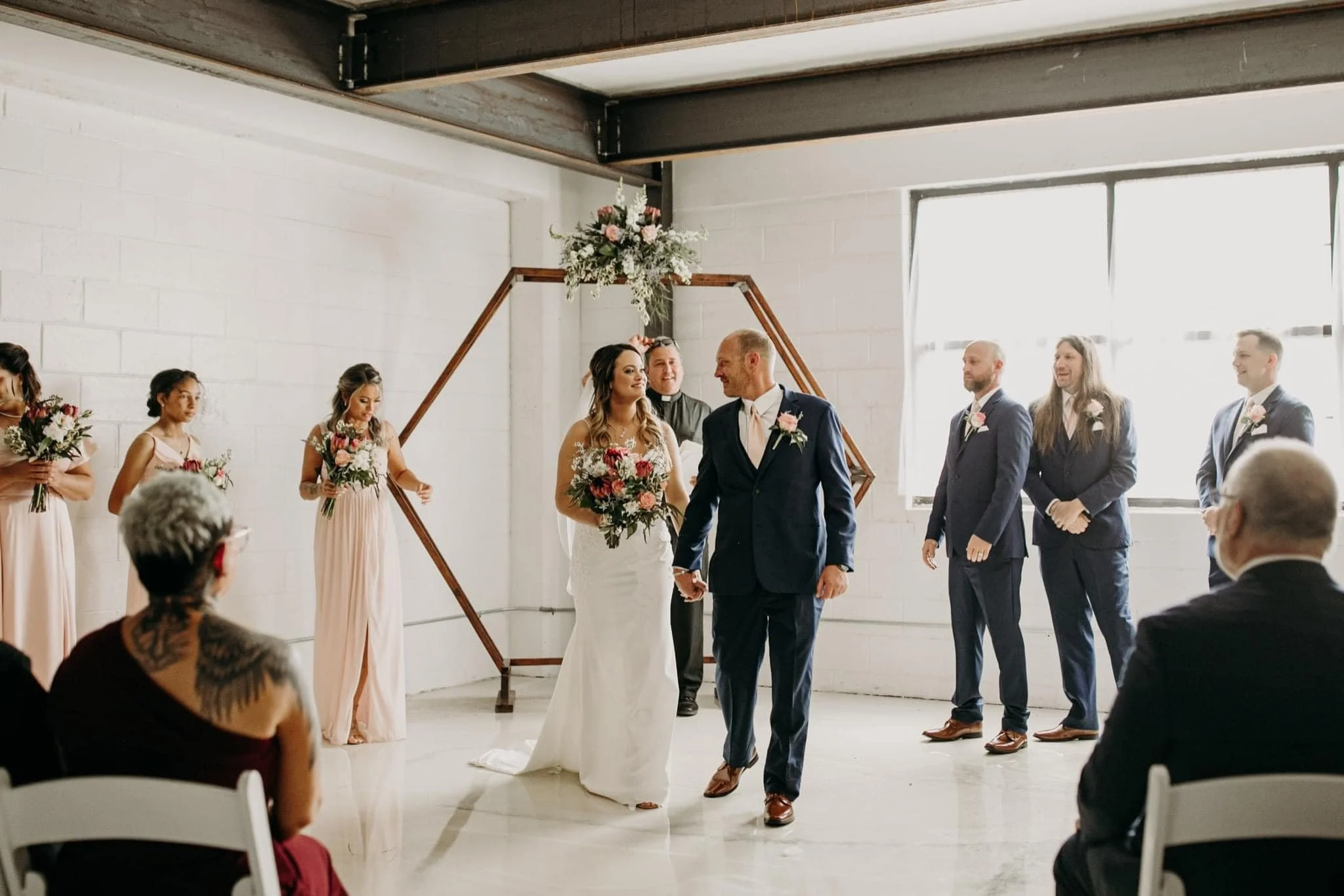 Bride and groom walking hand in hand down the aisle during their wedding ceremony, surrounded by bridesmaids and groomsmen, with guests seated in front. Dream Day Event Rentals, Vintage and Modern Decor and Floral Rentals, Freeport Illnois.