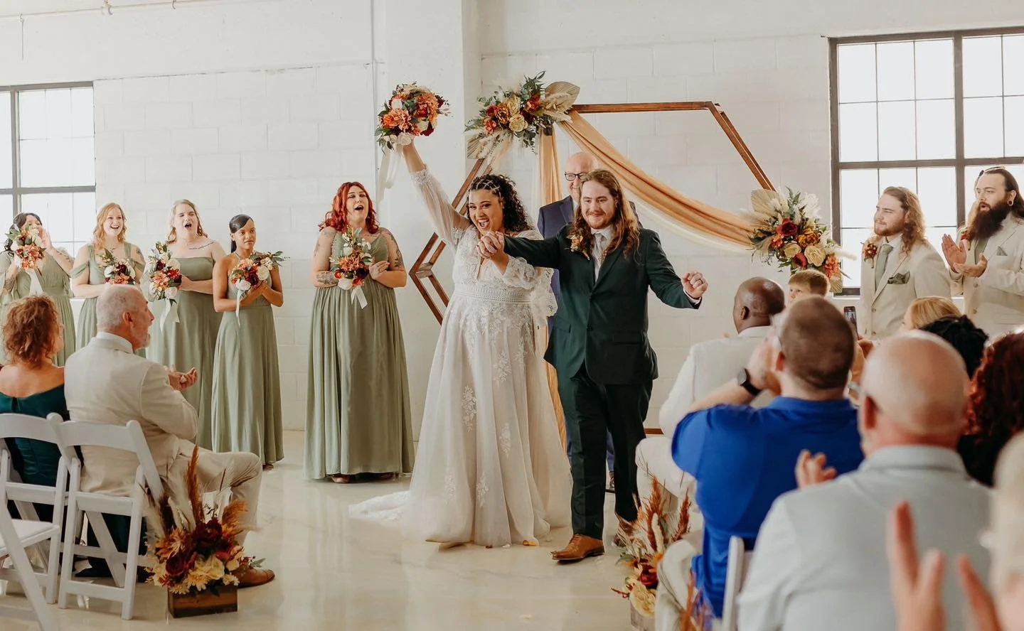 A wedding ceremony with a bride and groom crossing the finish line with a ribbon, surrounded by bridesmaids, groomsmen, and guests in a brightly lit, decorated venue with large windows and floral arrangements.